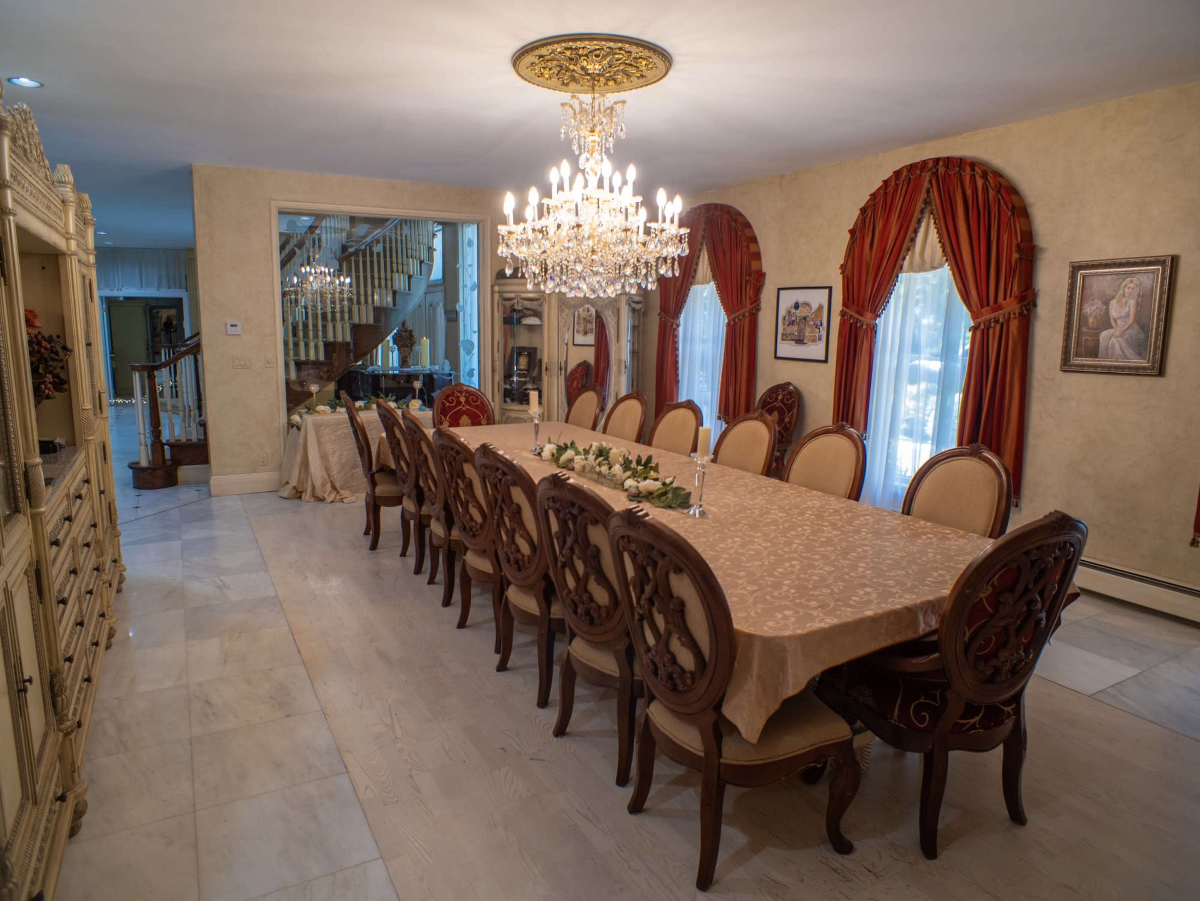 The image shows a well-furnished dining room featuring an elongated table surrounded by ornate chairs, with a chandelier above and decorative curtains at the windows.