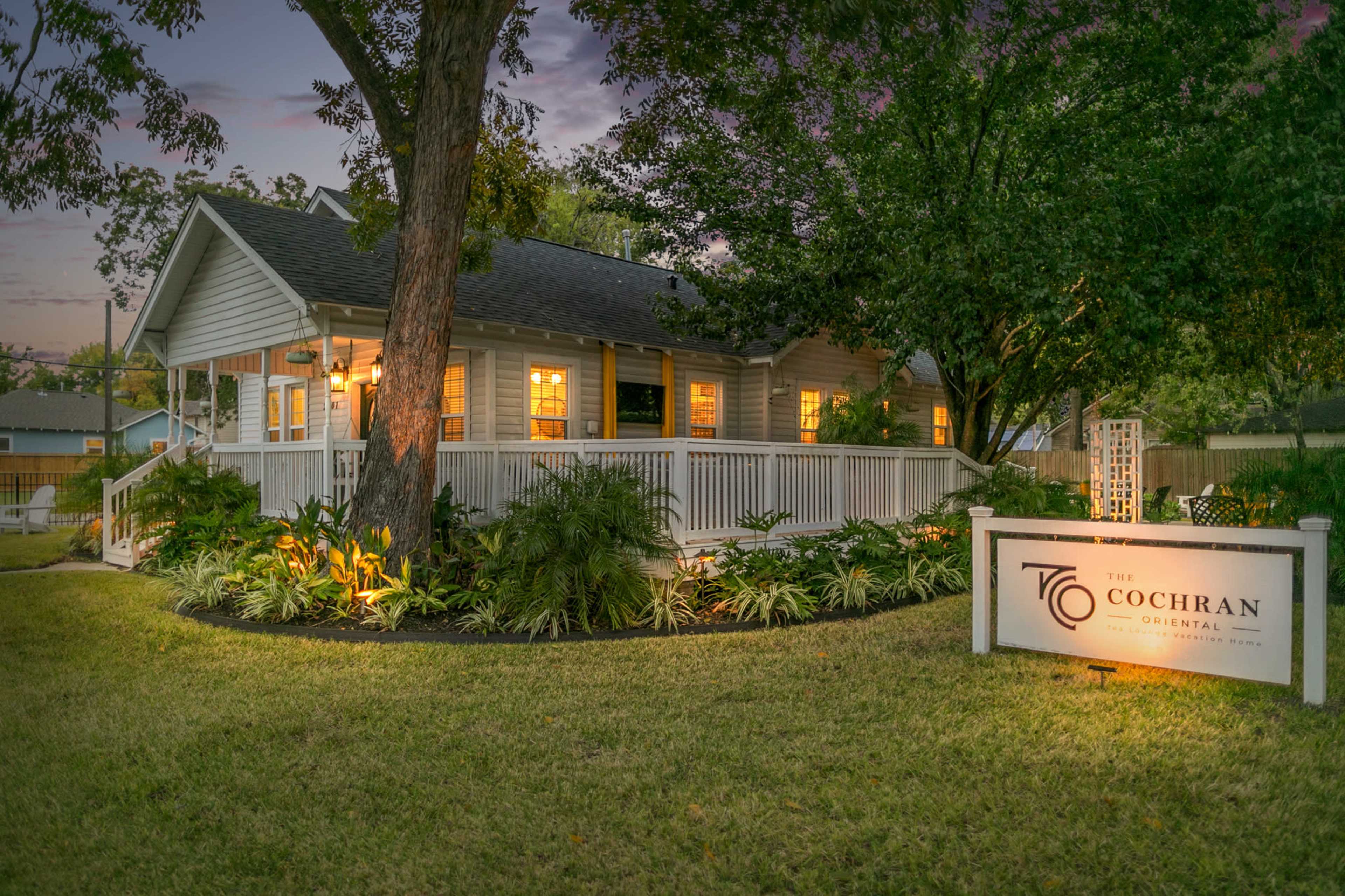 A white house with a porch is surrounded by lush greenery and illuminated by lights, featuring a sign that reads "The Cochran."