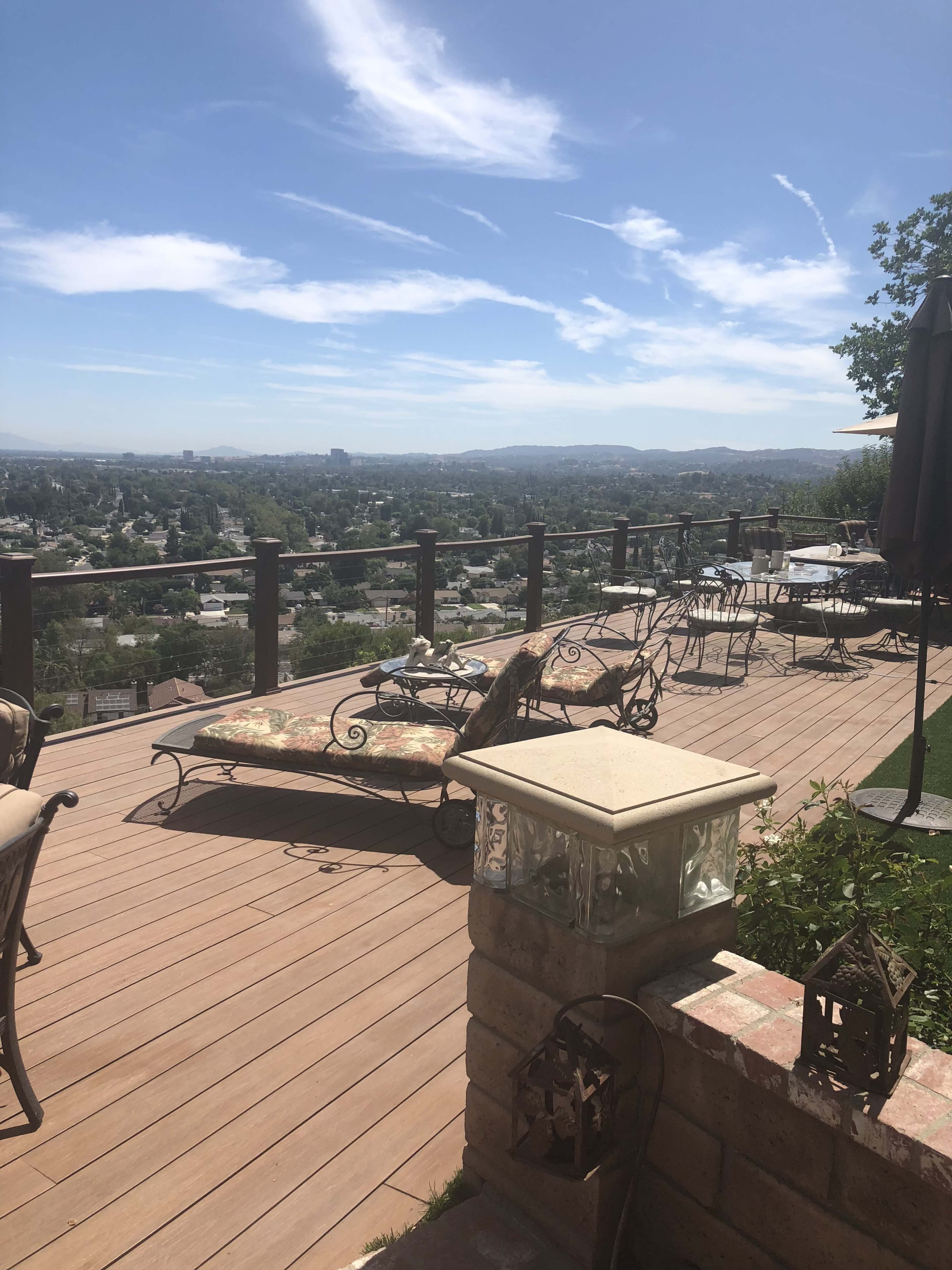 A wooden deck with lounge chairs and tables overlooking a suburban landscape under a clear blue sky.