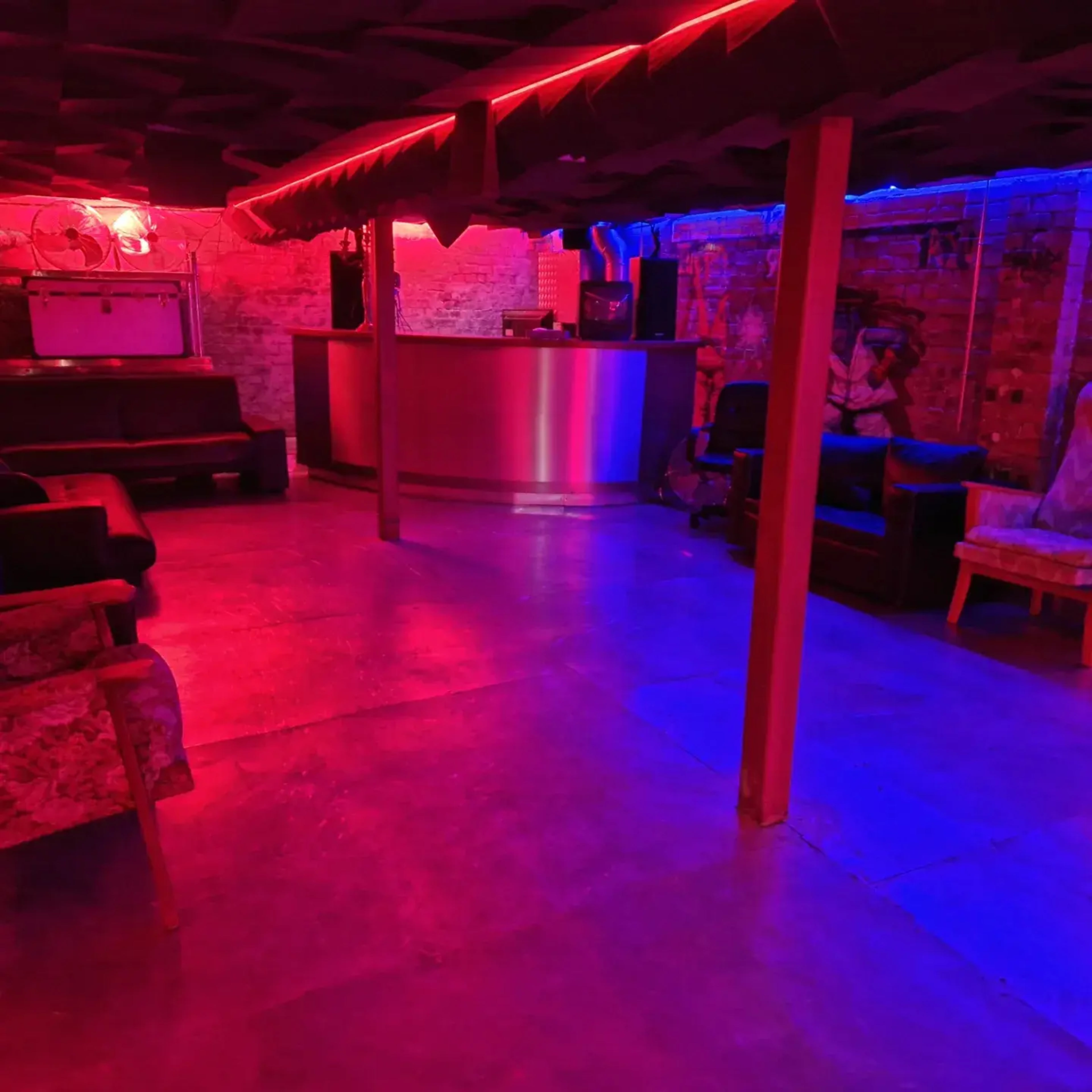 A dimly lit basement area with colorful neon lighting, featuring a bar counter and various seating options.