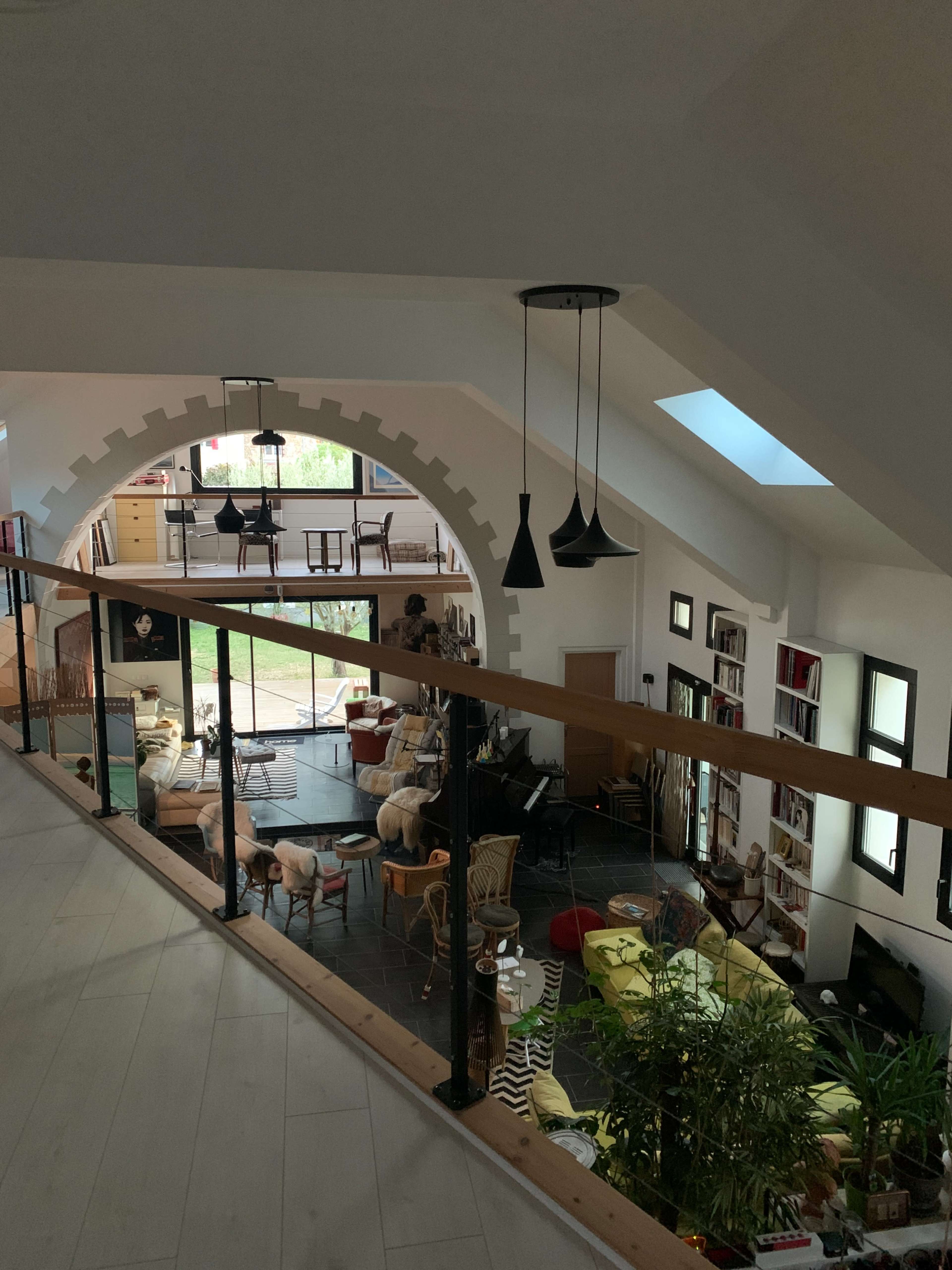 A spacious, modern interior features a balcony overlooking a lower living area filled with various furniture and plants, illuminated by natural light streaming through large windows.