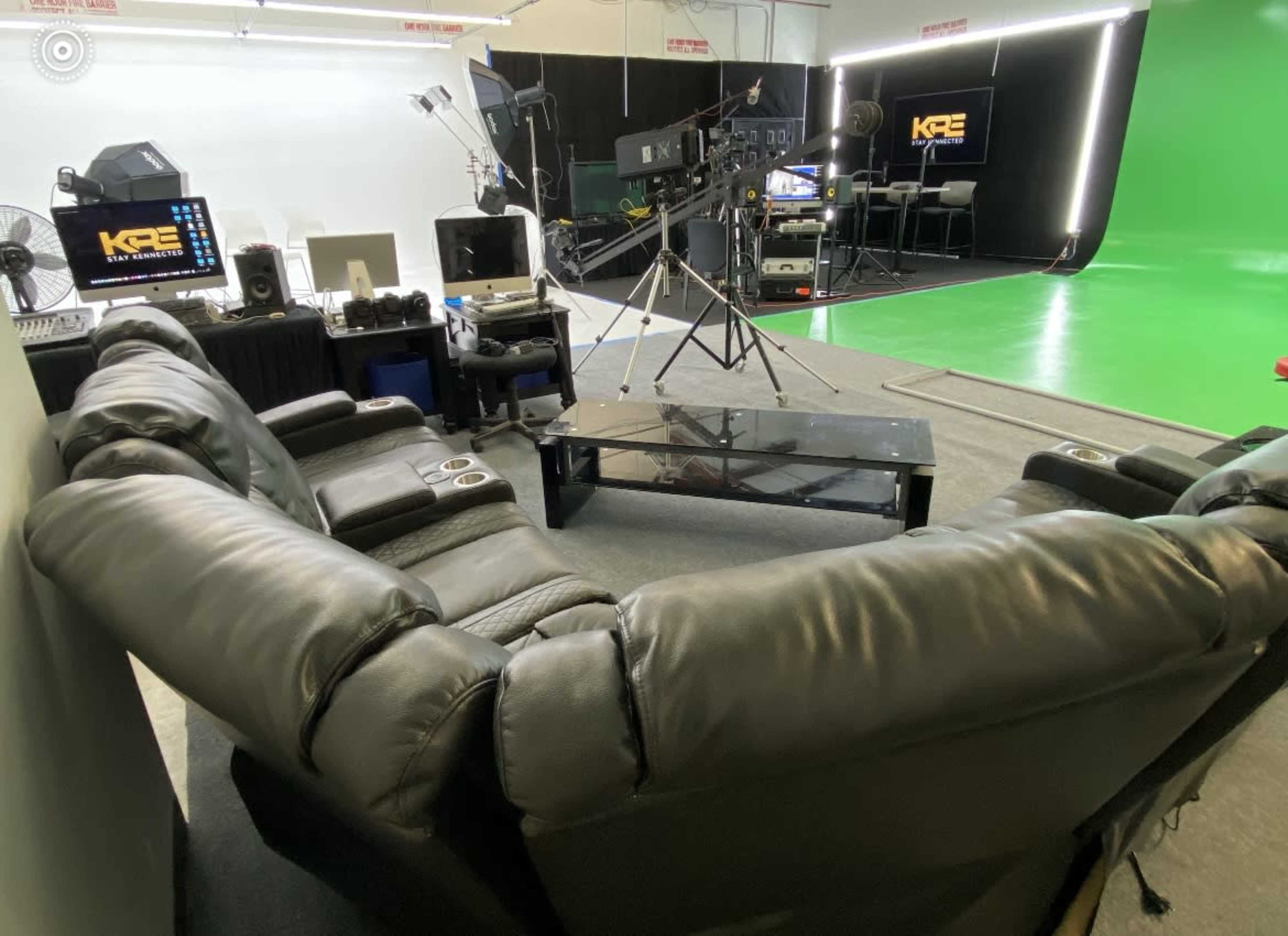 A circular black leather couch positioned around a glass coffee table in a studio setting with a green screen backdrop and professional equipment.
