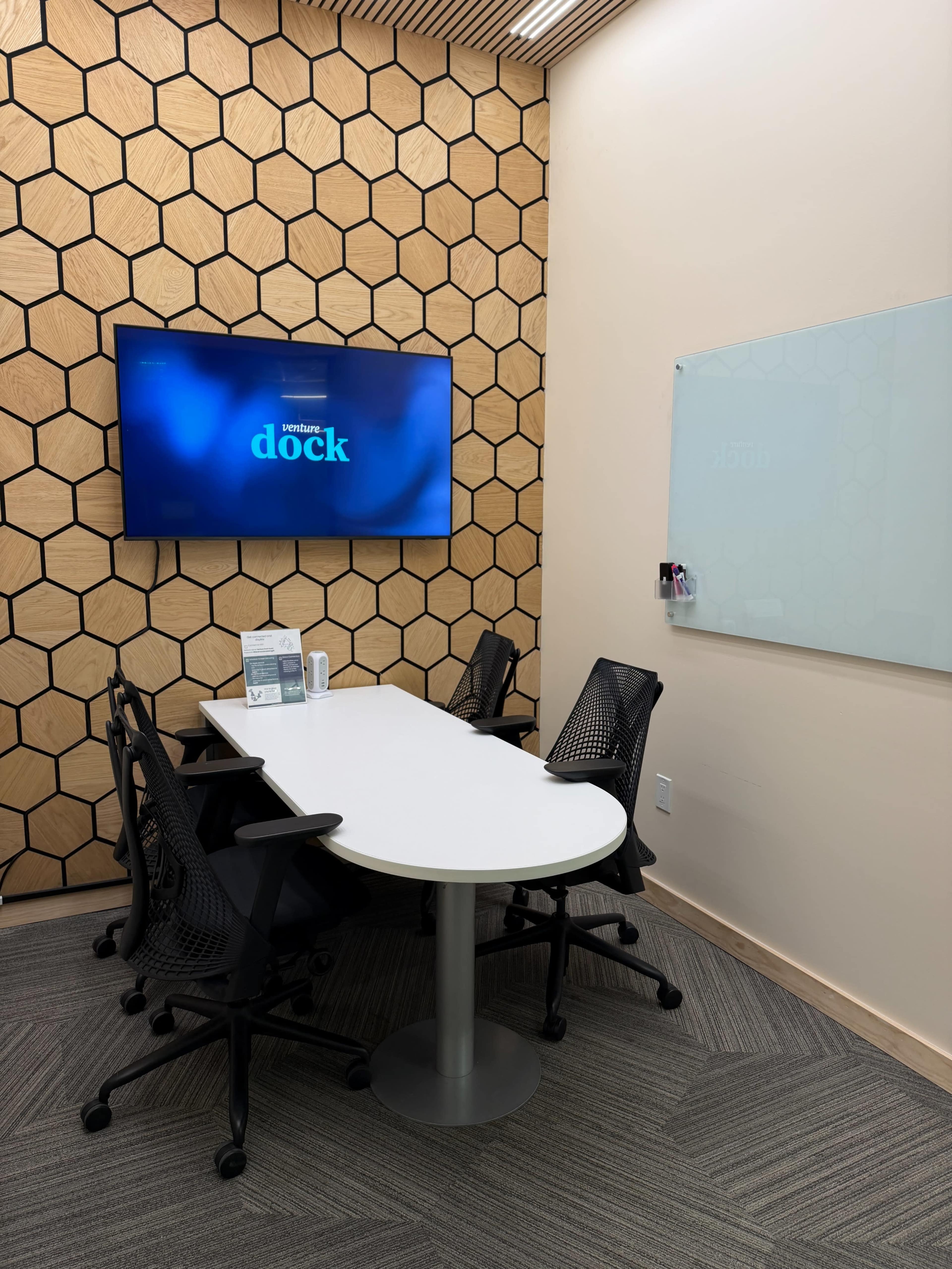 The image shows a small conference room with a white table, four black mesh chairs, a large screen mounted on the wall displaying “dock,” and a glass whiteboard.