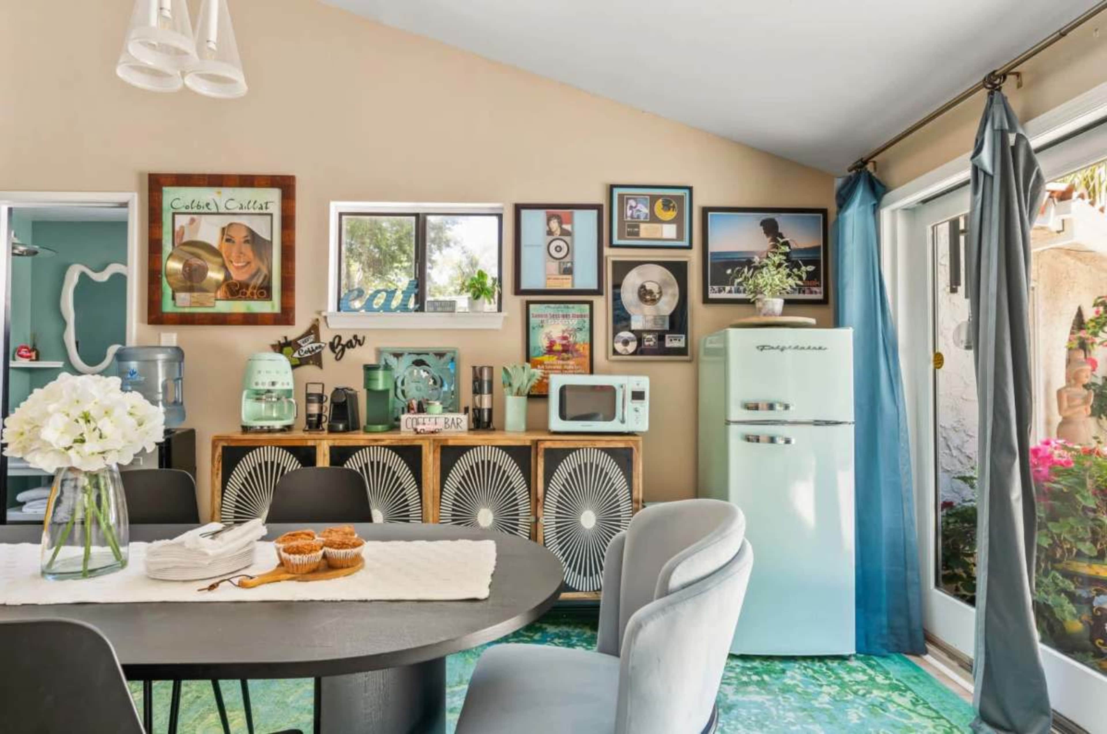 The image shows a brightly decorated kitchen with a round table, mint-colored appliances, and a collage of framed photos and records on the wall.