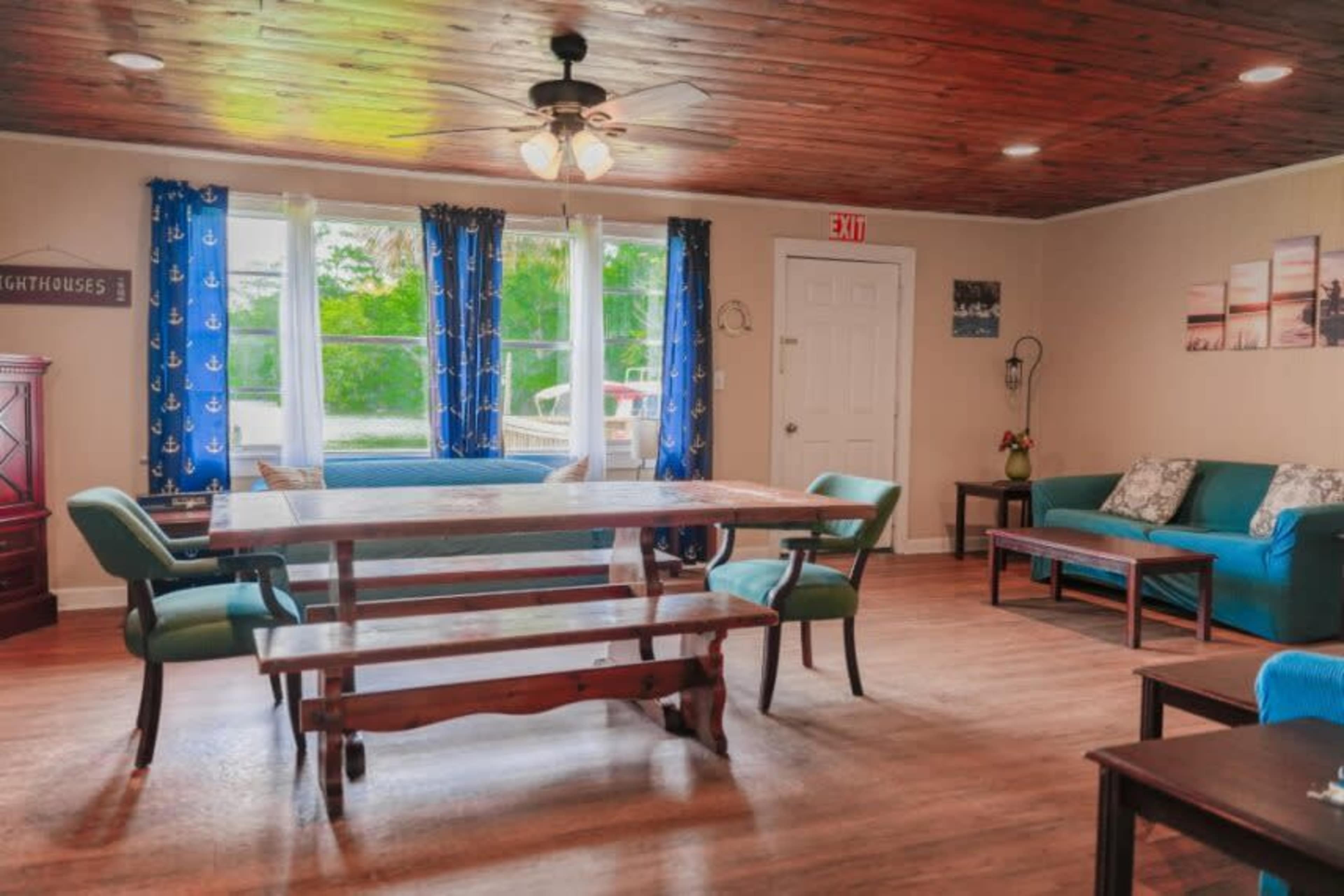 A room with a wooden dining table and benches, surrounded by green and blue furniture, large windows, and nautical-themed curtains.