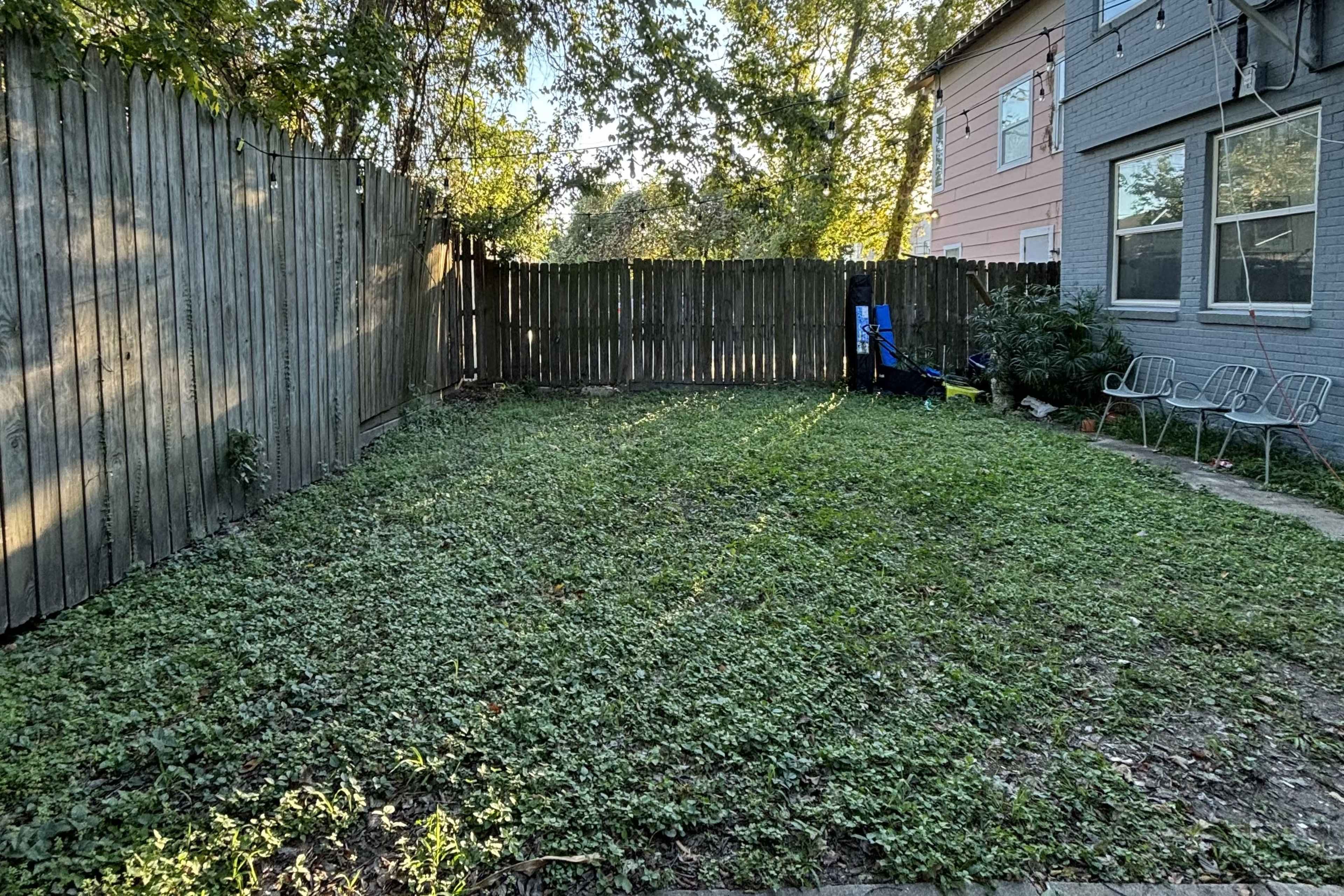 A fenced backyard with sparse grass and a few outdoor chairs near a house.