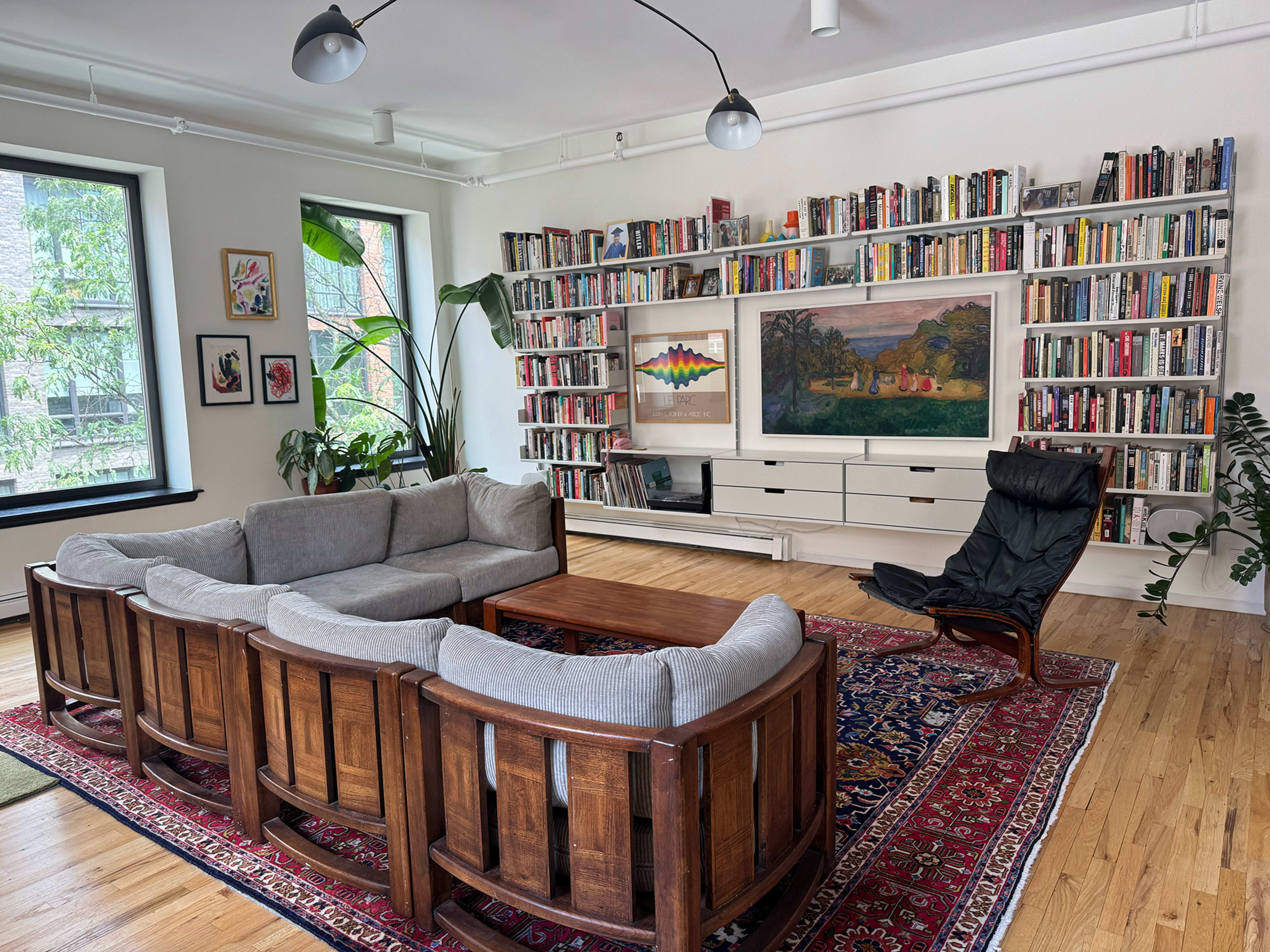 The image shows a modern living room featuring a circular wooden seating arrangement, a coffee table in the center, a large bookshelf filled with books, and a framed painting on the wall.