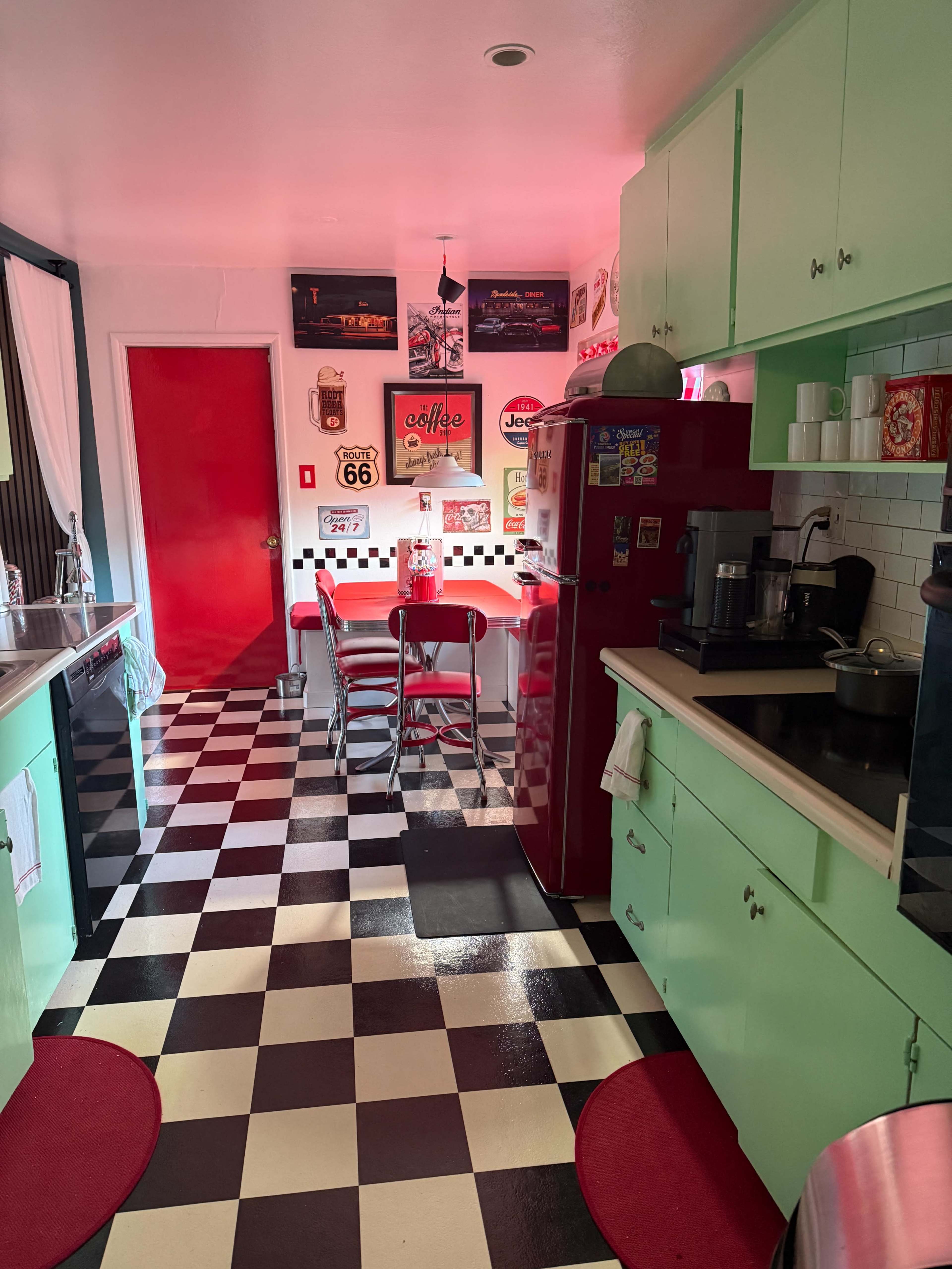 The image shows a retro-style kitchen with mint green cabinets, a red refrigerator, and a black-and-white checkered floor, featuring a small dining area with red stools.