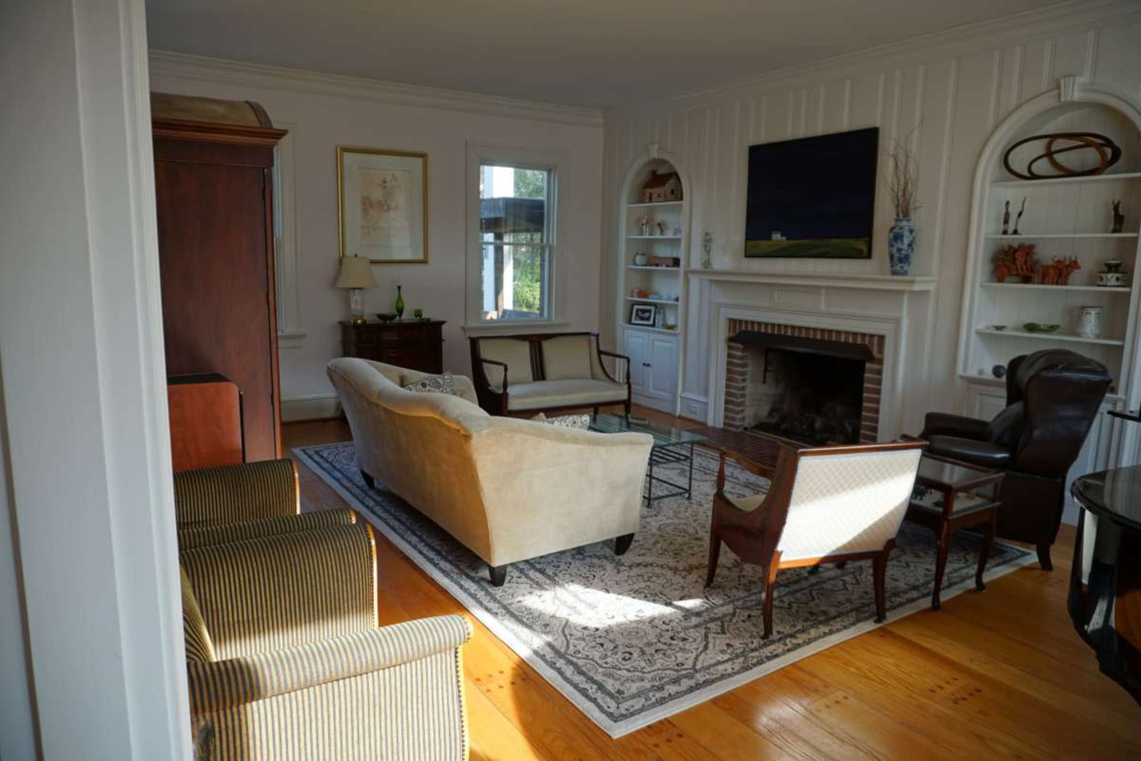A bright living room features a sofa, armchairs, and a coffee table arranged around a fireplace, with bookshelves and a television on the wall.