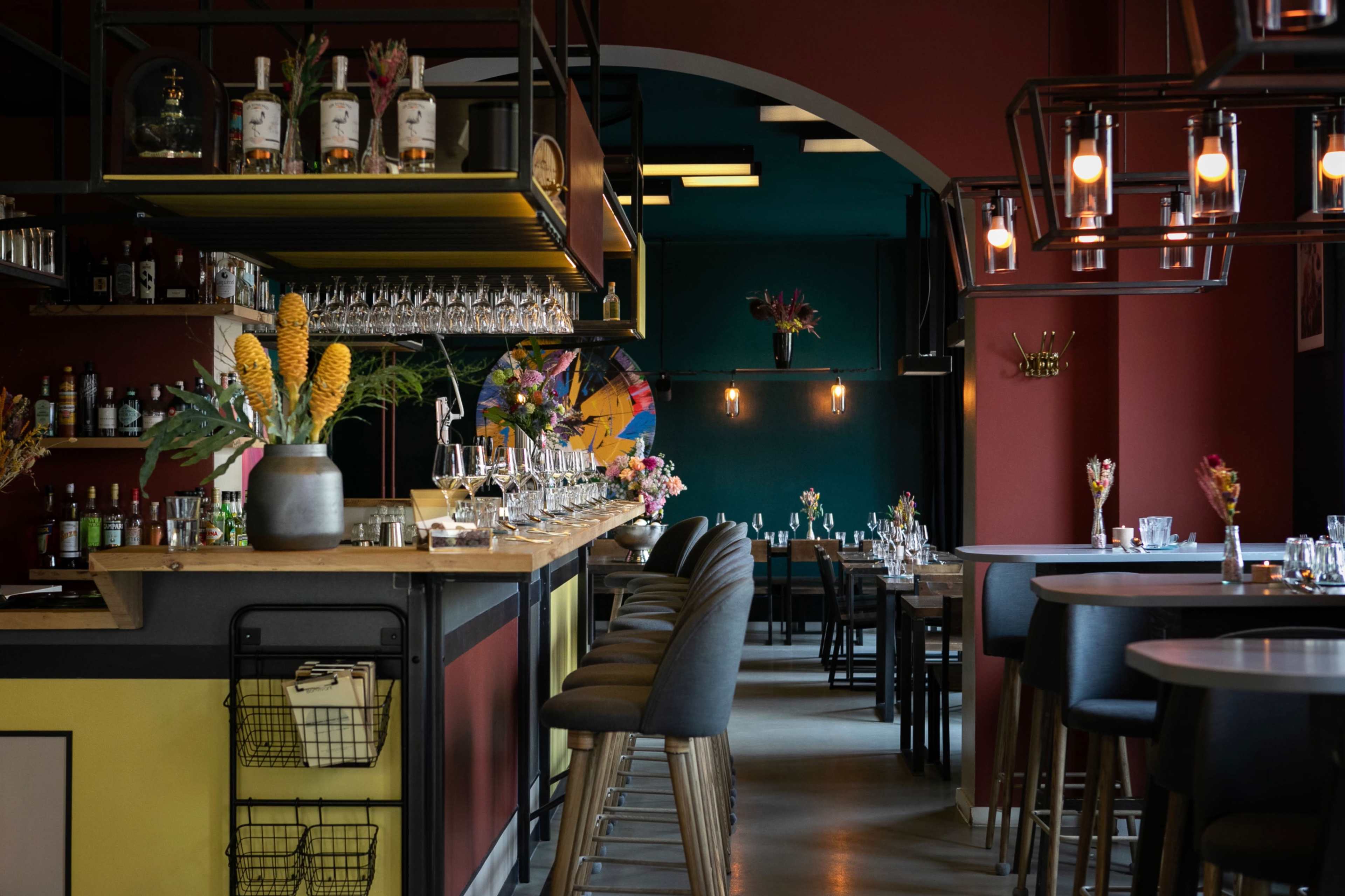 The image depicts a modern restaurant interior featuring a bar with colorful decor, high stools, and floral arrangements on tables.