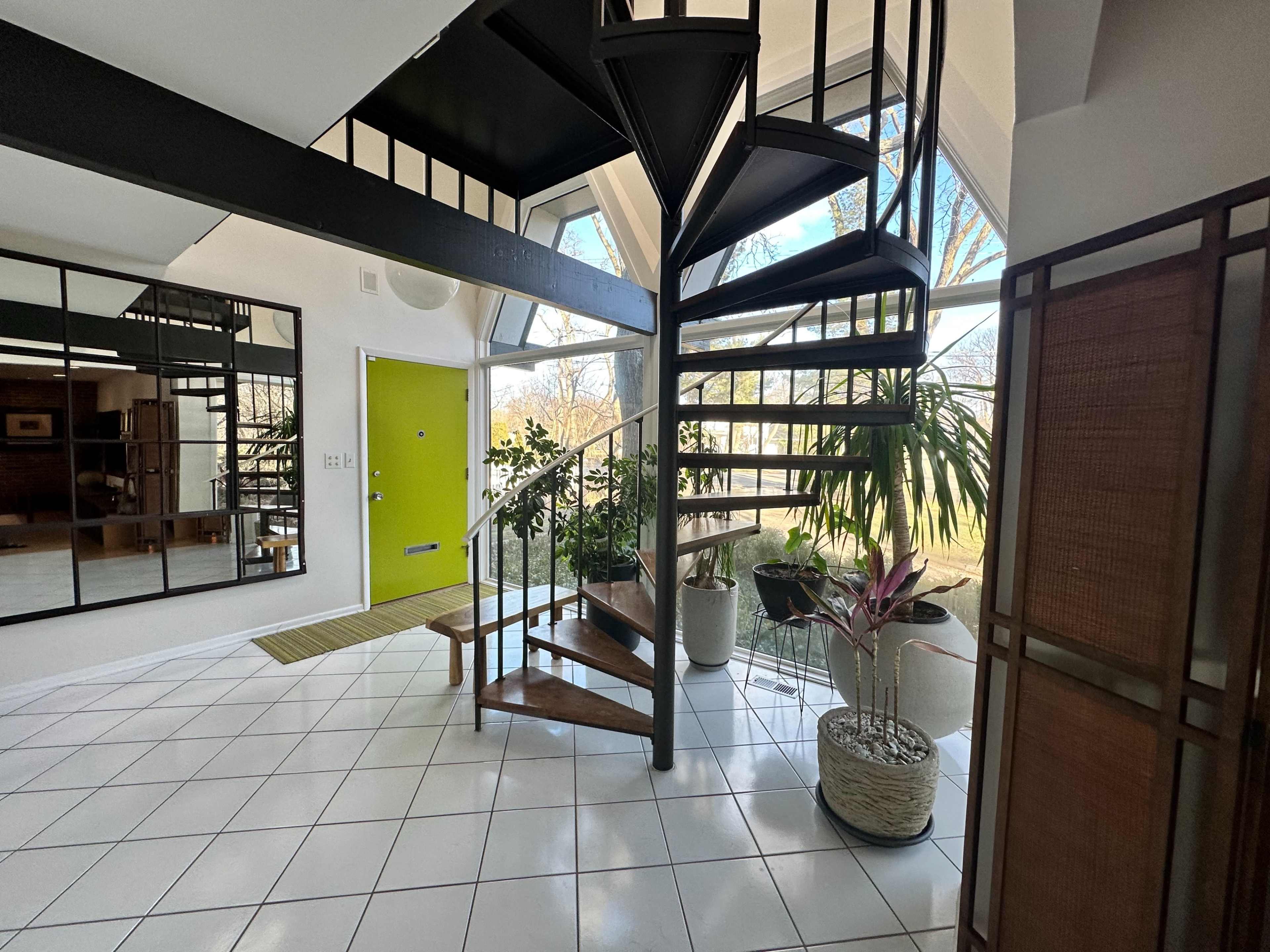 A modern entryway with a spiral staircase, large windows, and potted plants arranged around a tiled floor.