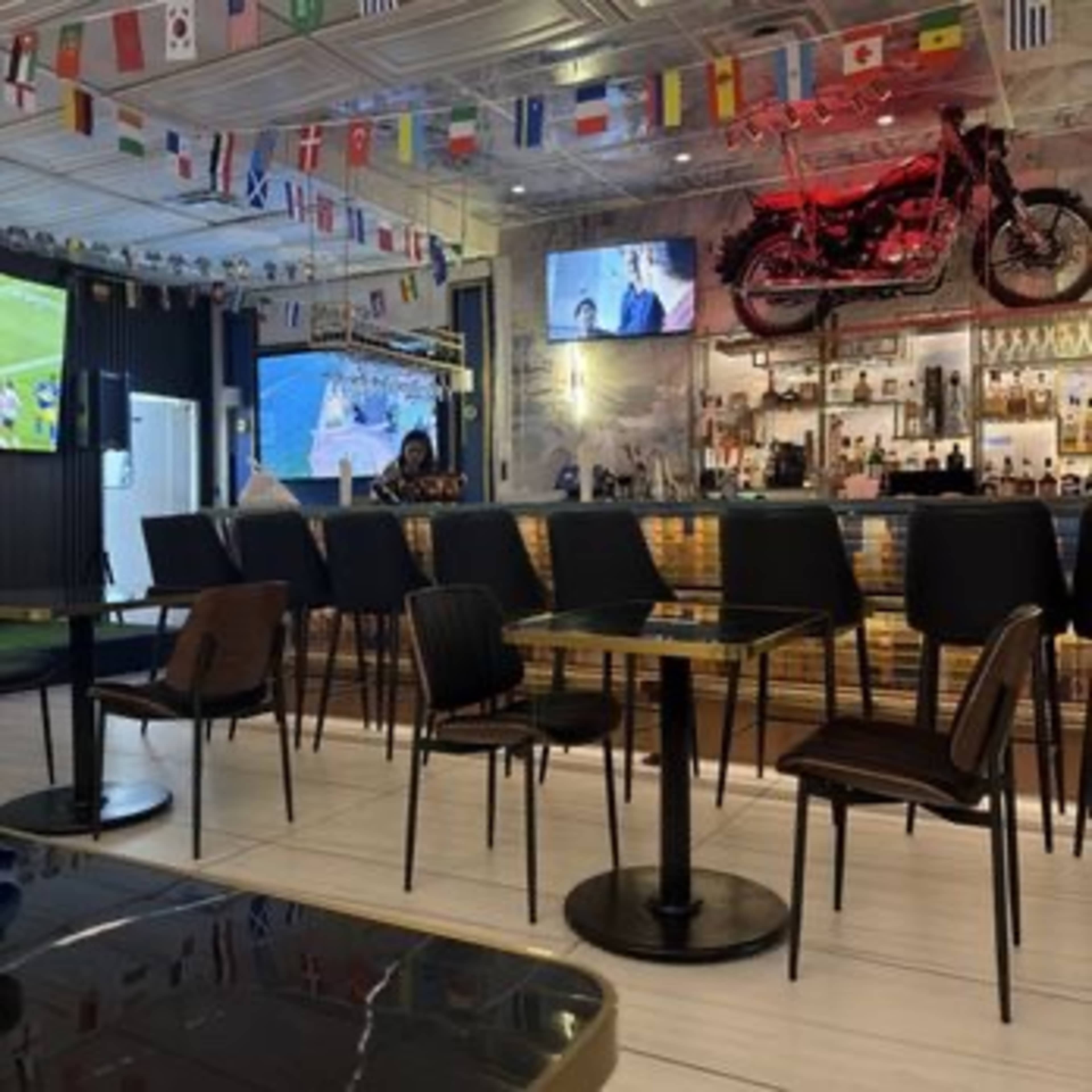 The image shows a modern bar or restaurant with seating and a display showcasing international flags, a motorcycle on the wall, and a large screen broadcasting a sports event.