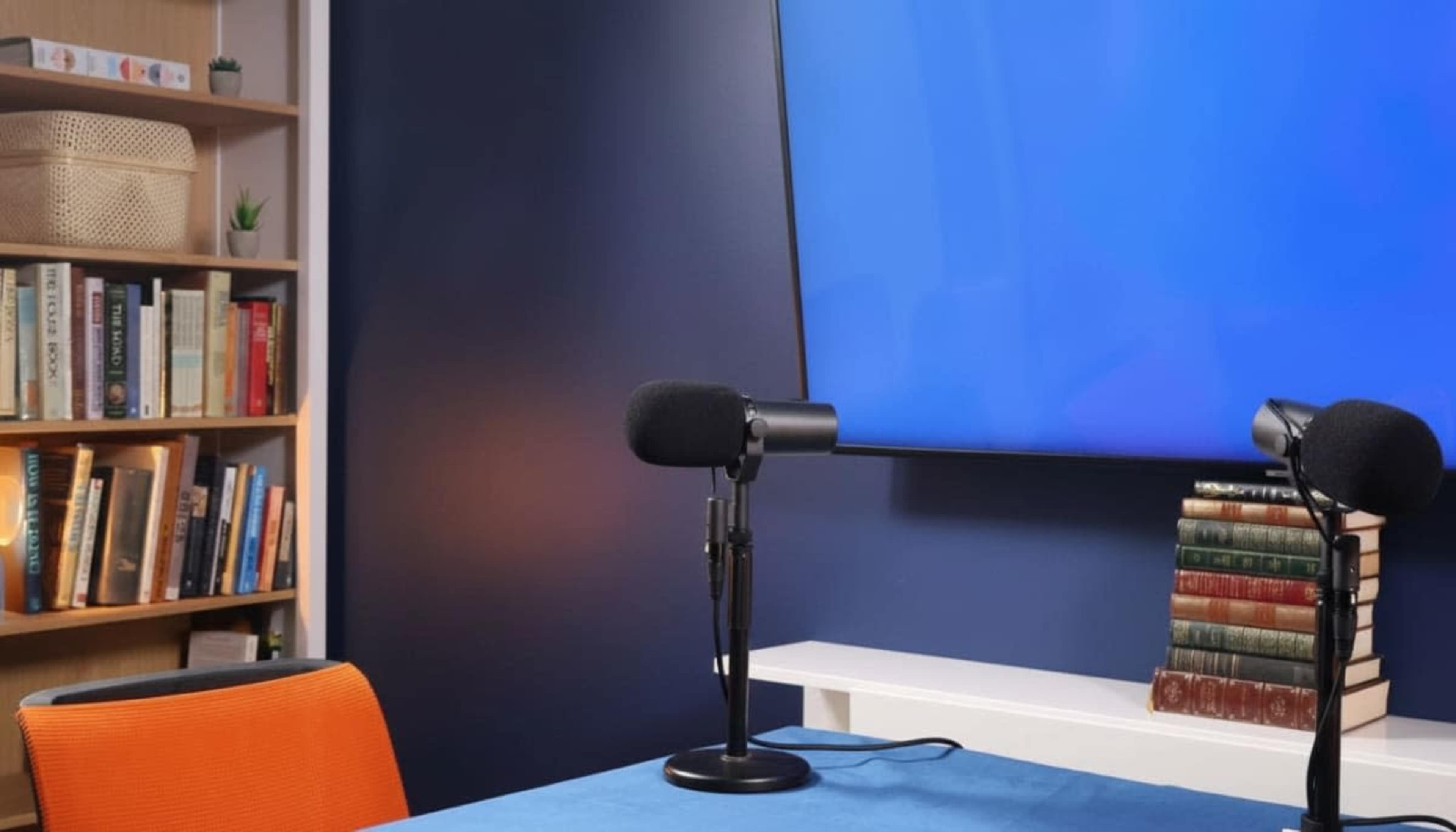 A well-organized podcast studio features two microphones on a table, a blue backdrop, and a bookshelf filled with books.