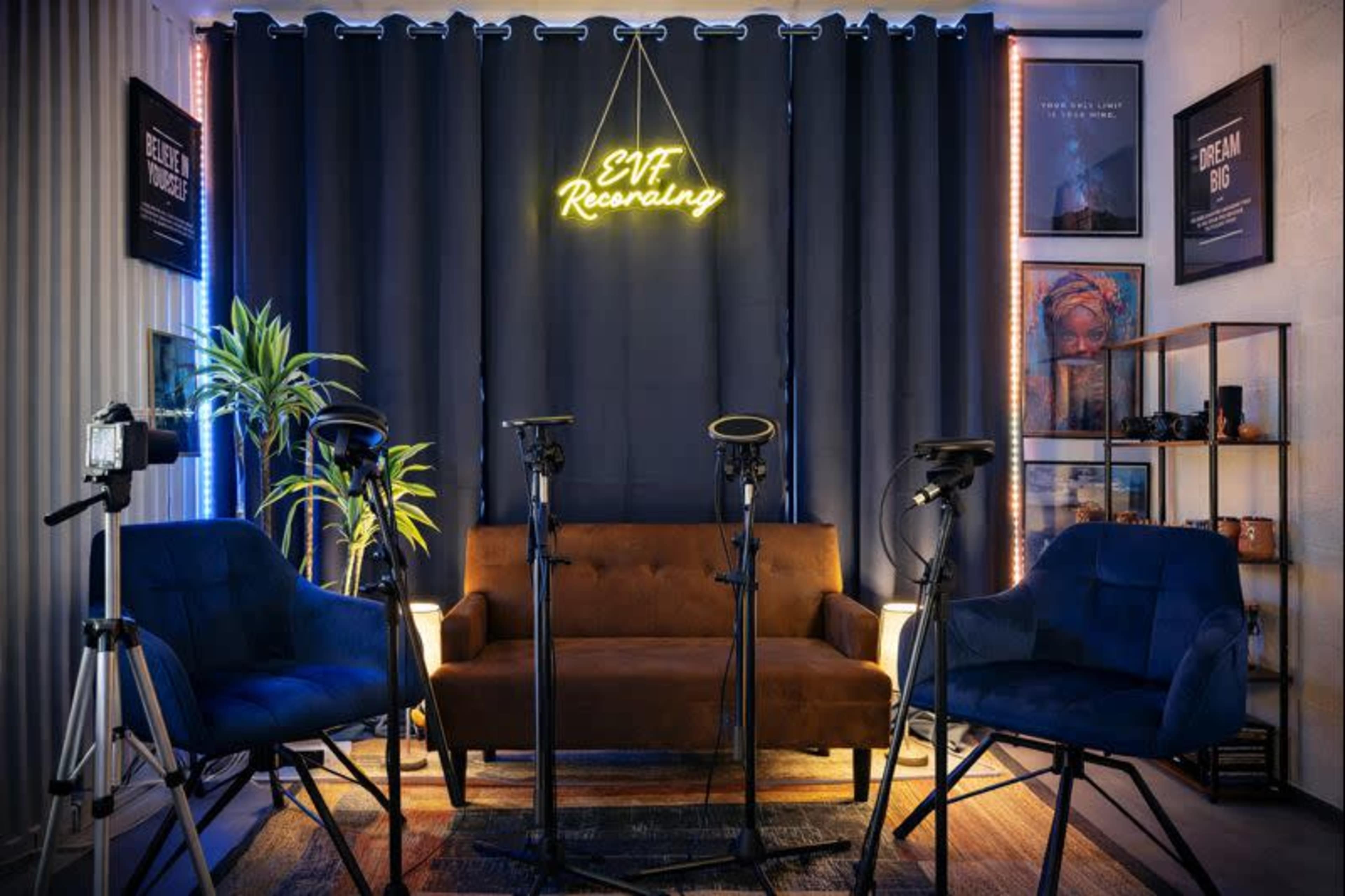 The image shows a cozy recording studio setup with a brown sofa, four microphones on stands, large black curtains, and soft lighting.
