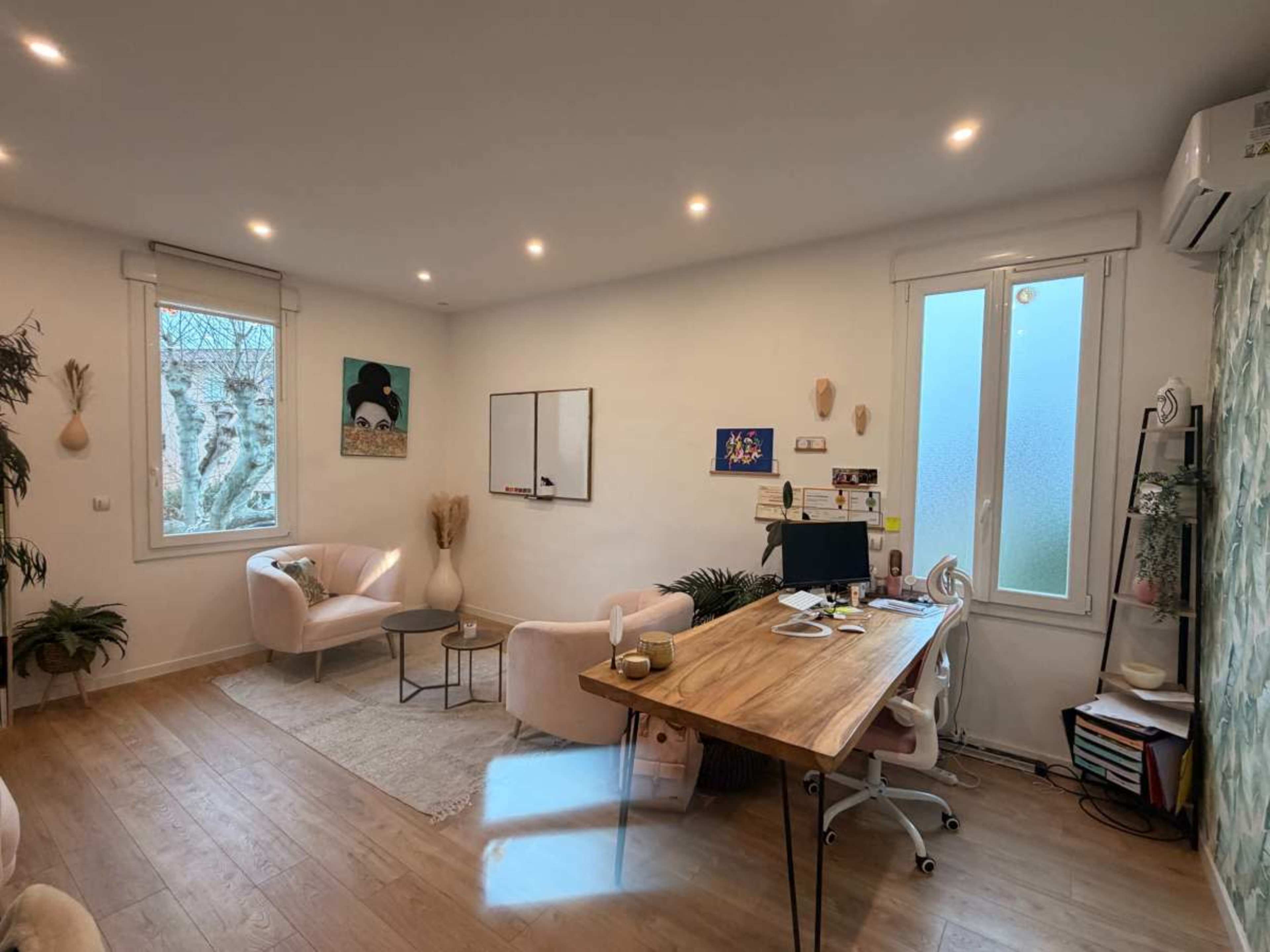 The image shows a modern office space with a wooden desk, a computer, two pink chairs, and large windows allowing natural light to enter.