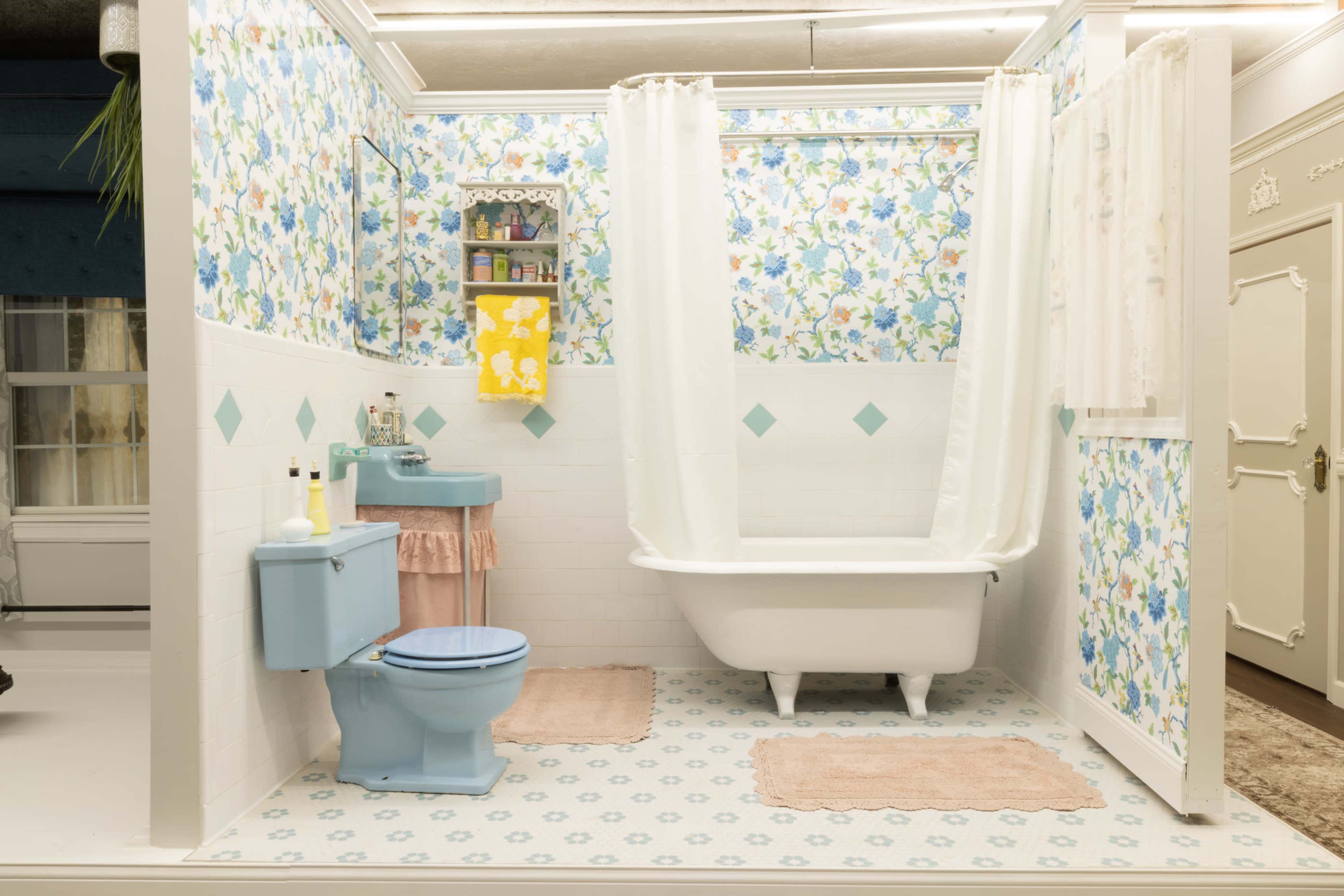 A bright bathroom features floral wallpaper, a blue toilet, a pink vanity with a sink, and a white bathtub enclosed by a shower curtain.
