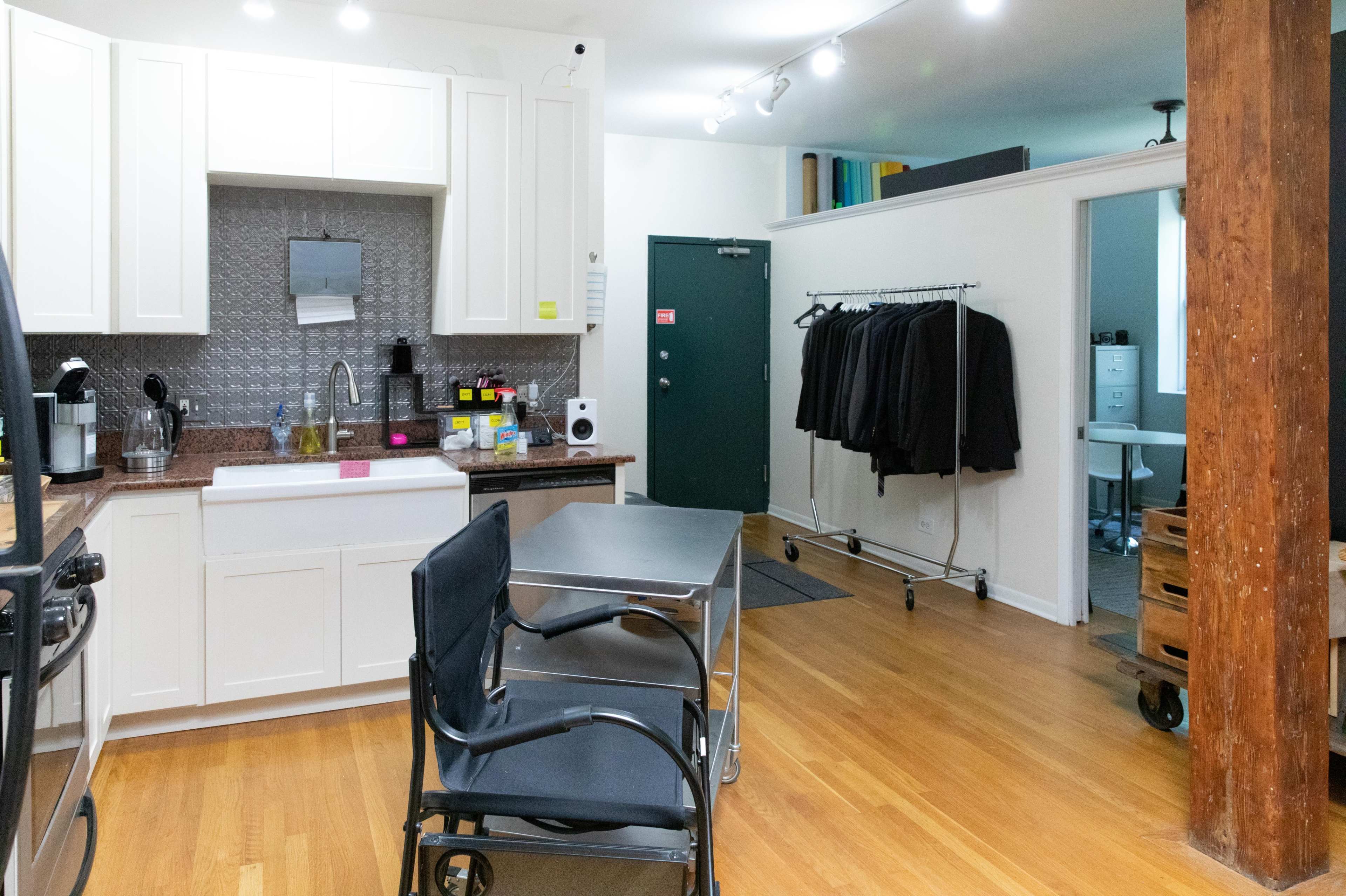 The image shows a modern kitchen space with a dining table and chairs, a clothing rack with jackets, and two doors leading to other rooms.