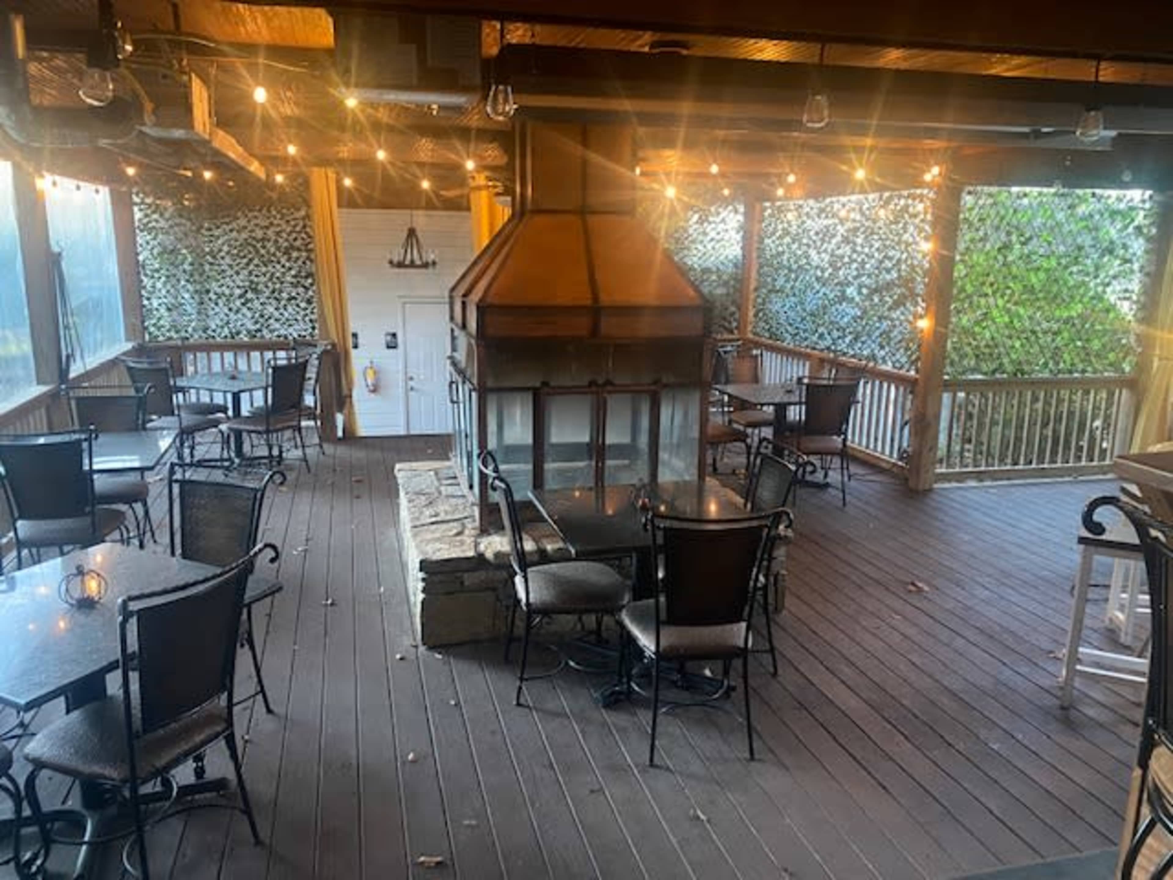A spacious dining area with tables and chairs arranged around a central fireplace, surrounded by walls covered in greenery and string lights overhead.
