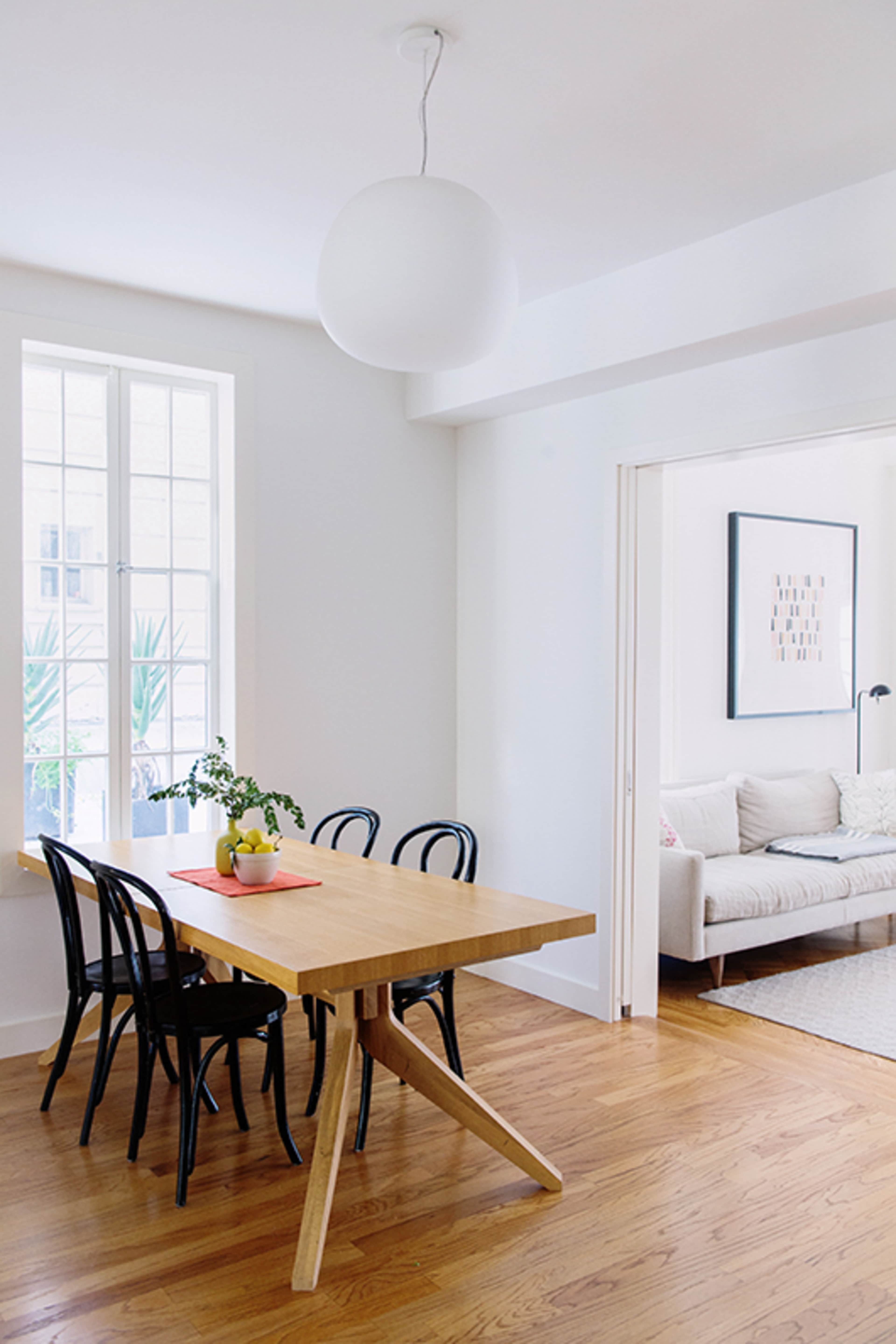 A simple dining area with a wooden table and four black chairs, accompanied by a light hanging above and a view of a living room through an adjacent doorway.