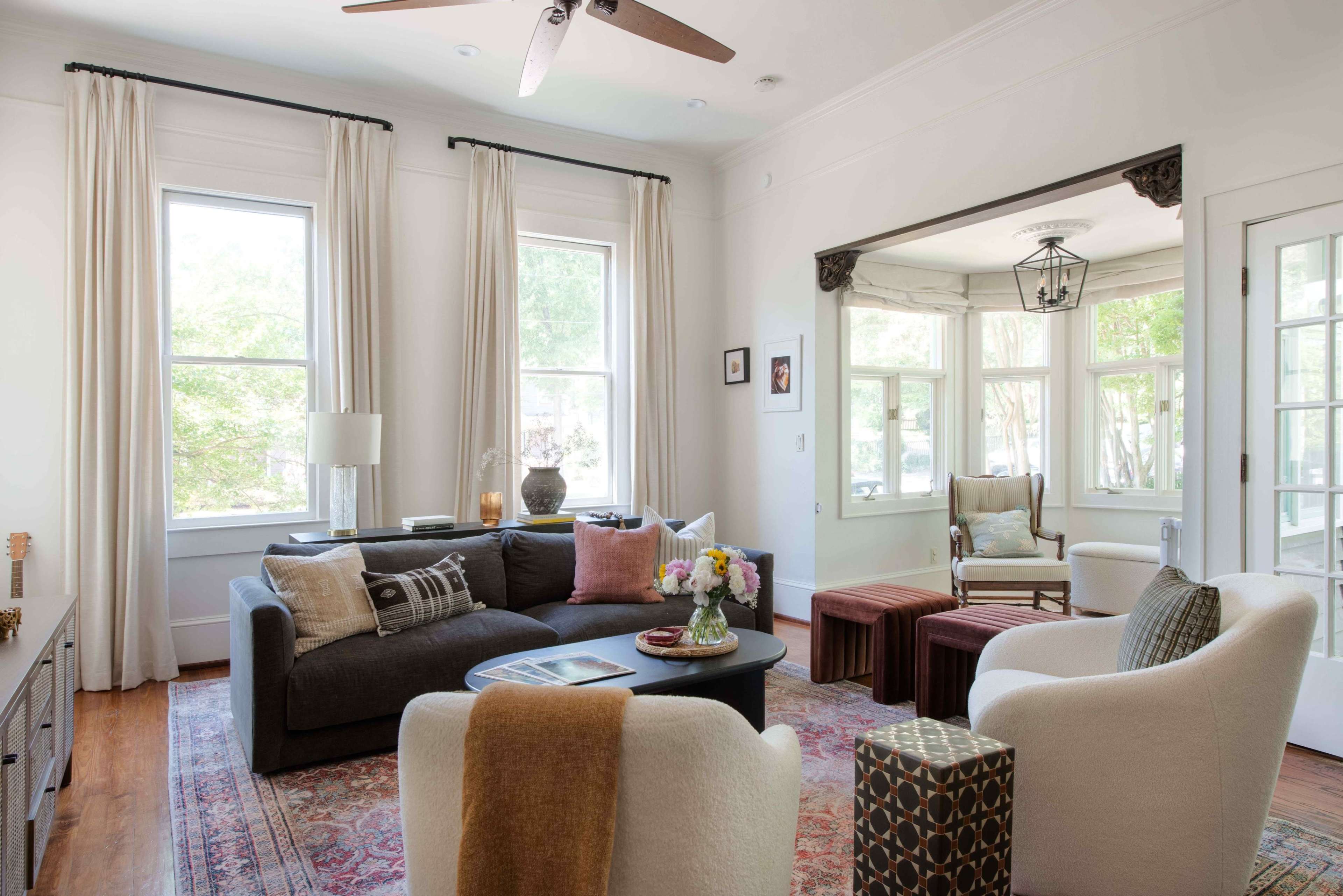 The living room features a dark sofa, white chairs, a coffee table, and large windows adorned with light curtains.