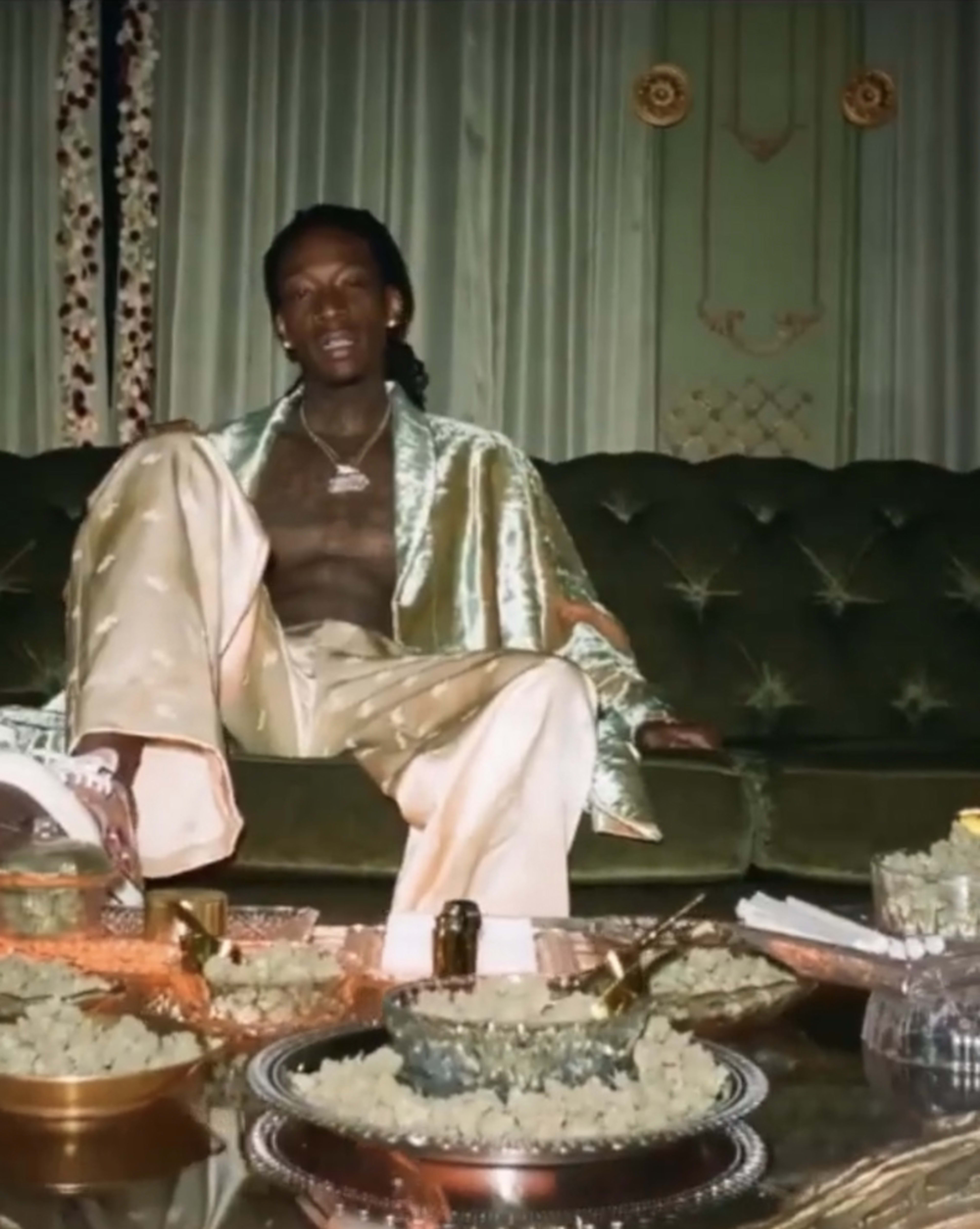 A man sits on a green velvet couch, wearing a light-colored outfit, with ornate dishes and decorations arranged on a low table in front of him.