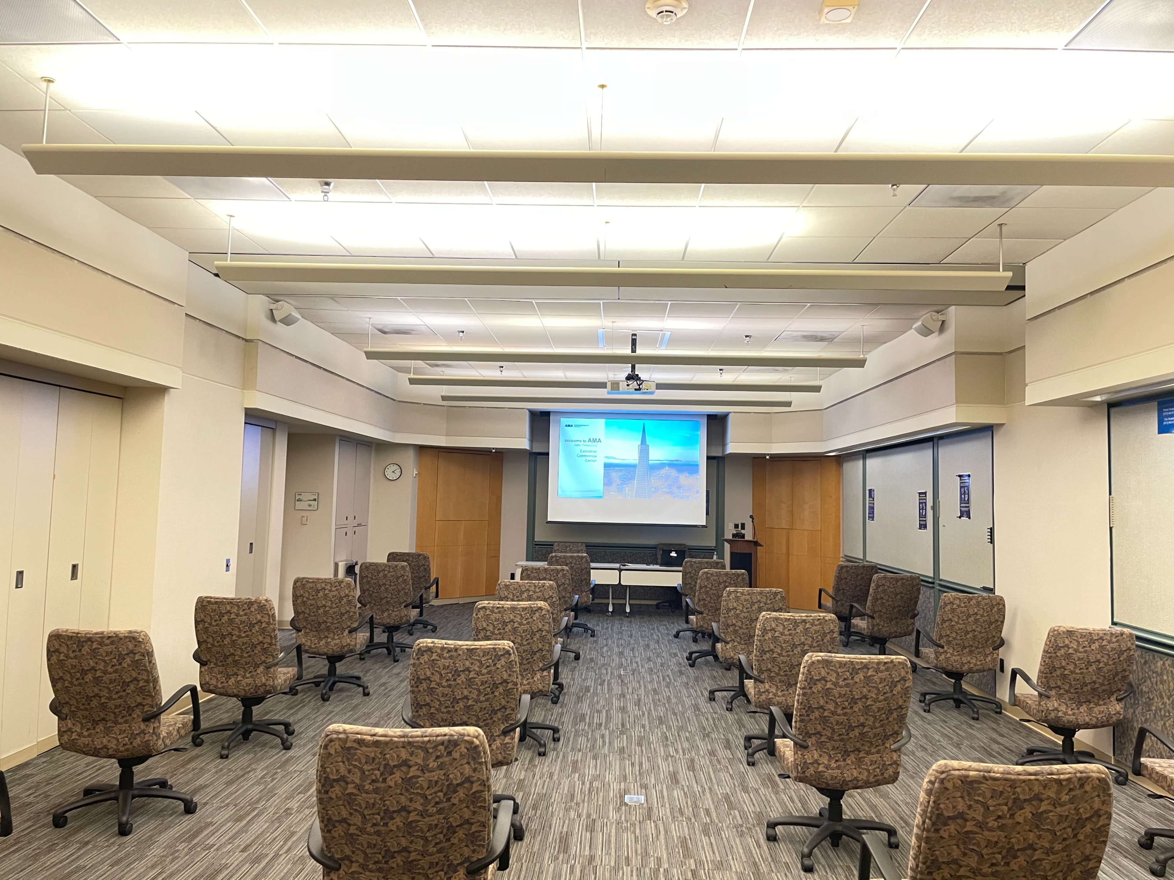A conference room is set up with rows of rolling chairs facing a projector screen.