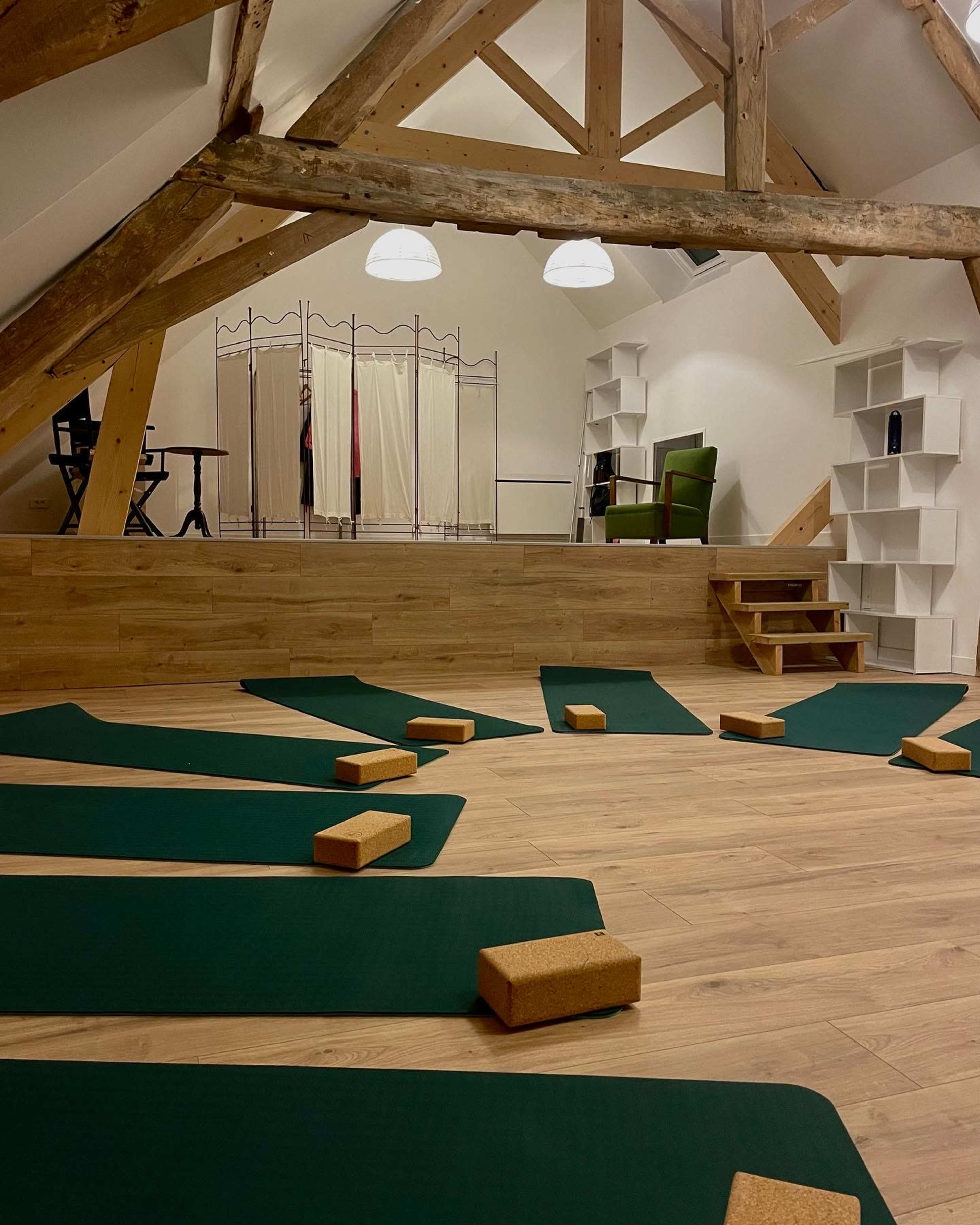 The image shows a spacious wooden-floored yoga studio with green mats laid out and cork blocks arranged on the floor, alongside a partitioned area and a few pieces of furniture.