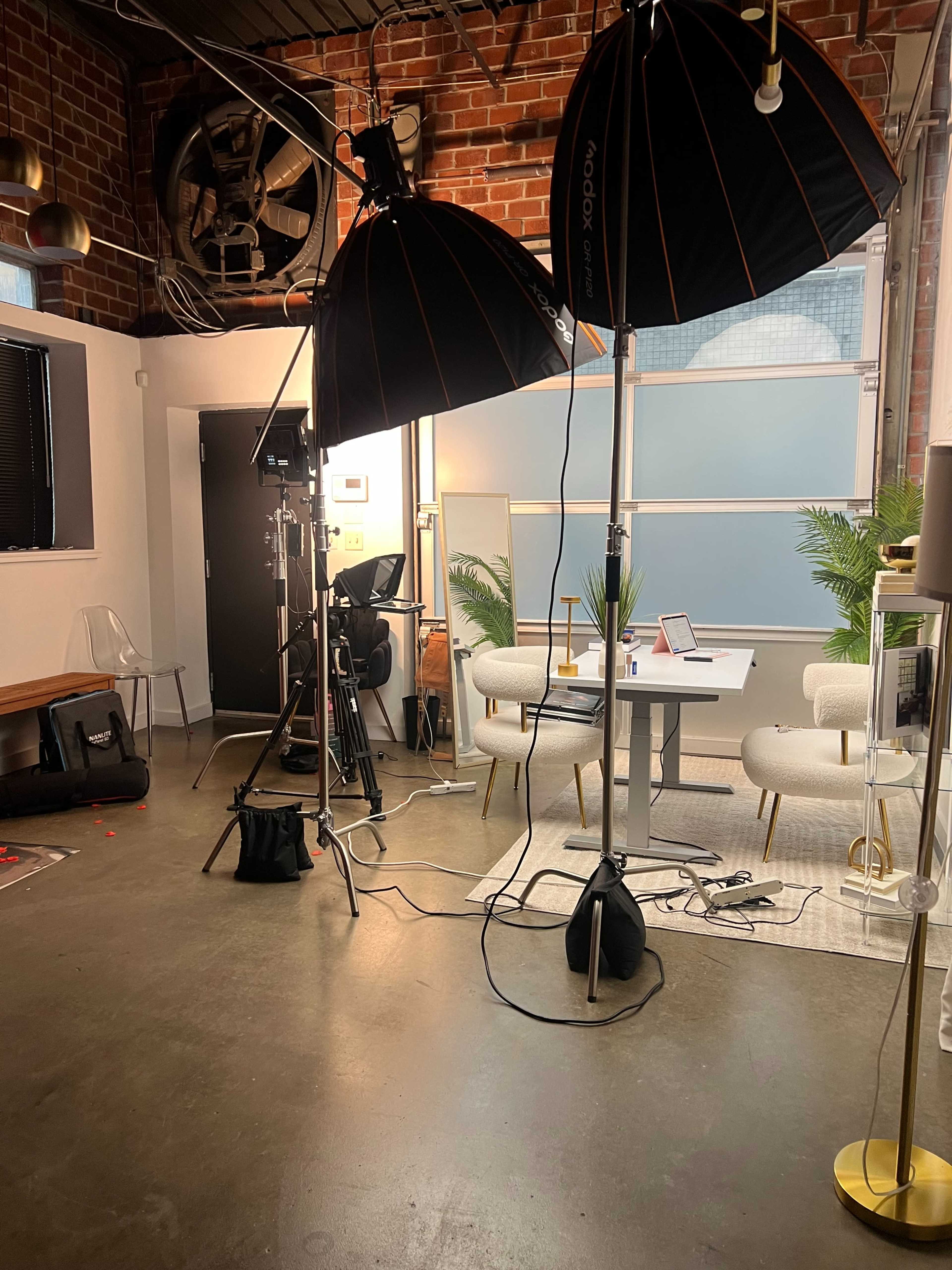 The image shows a well-lit studio setup with two large softbox lights, a camera on a tripod, and a modern seating area featuring a table and chairs against a brick wall.