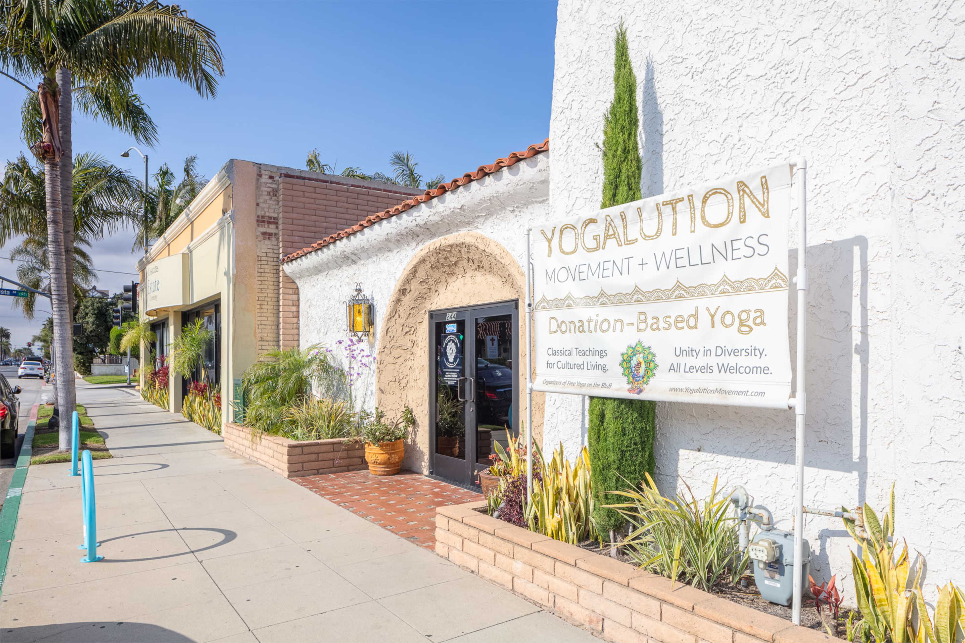 The image shows a yoga studio called "Yogalution Movement + Wellness," featuring a large sign indicating it offers donation-based yoga, alongside a shopping area with palm trees in the background.