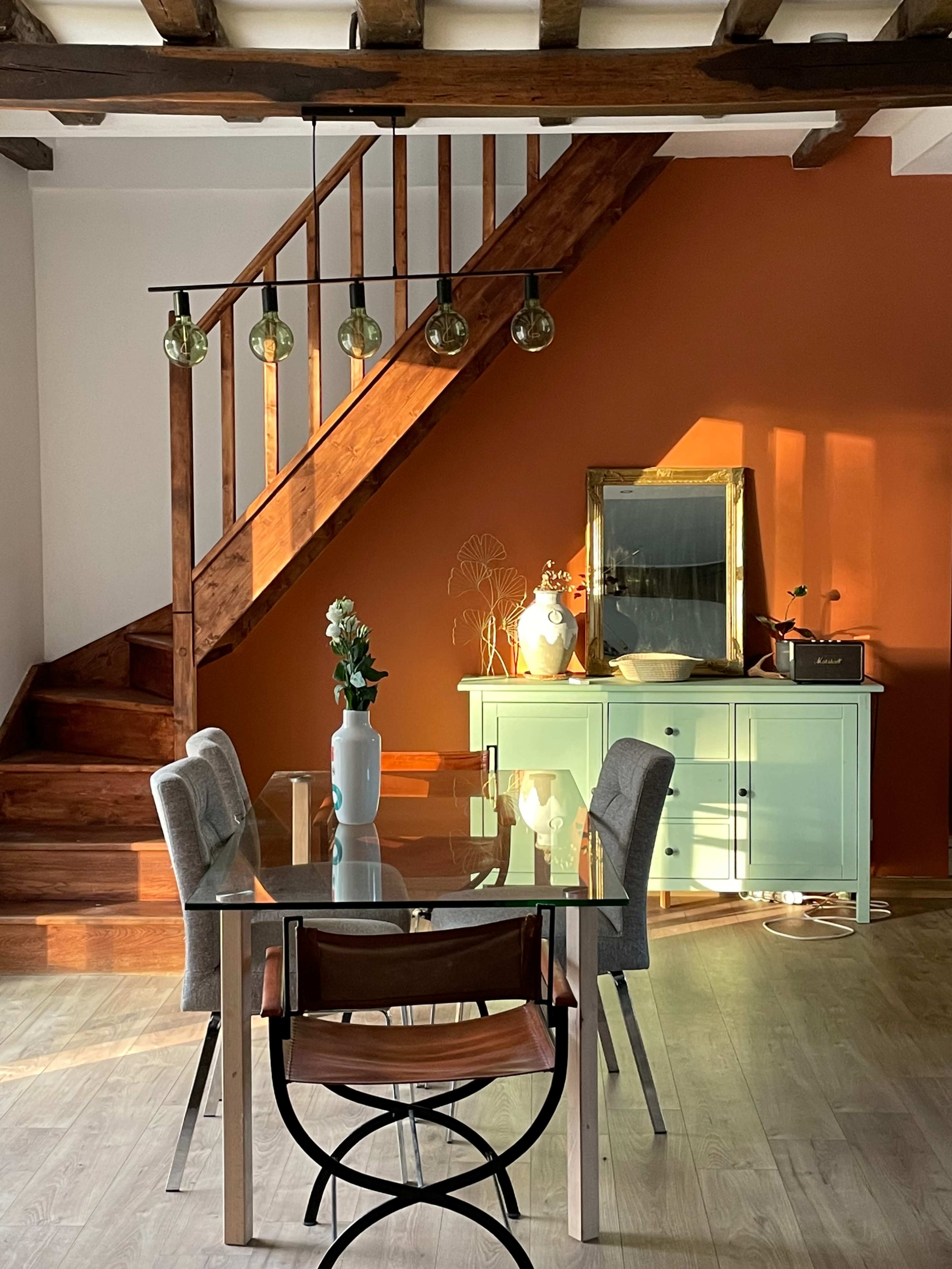 A dining area features a glass table with four chairs, a green sideboard, and a staircase illuminated by pendant lights.
