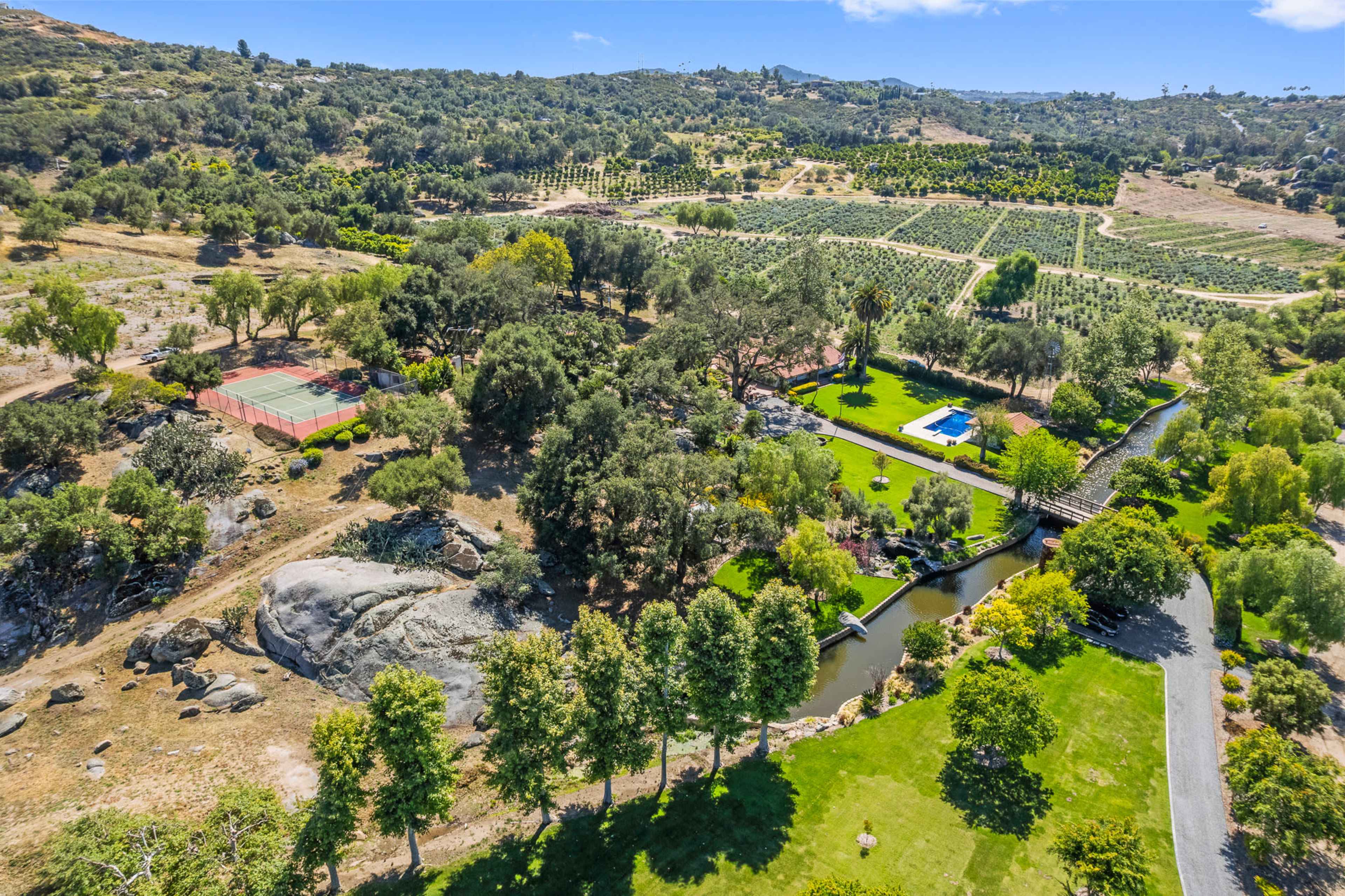 The image shows an aerial view of a sprawling landscape featuring a tennis court, a swimming pool, a winding creek, and neatly arranged rows of trees in a rural setting.