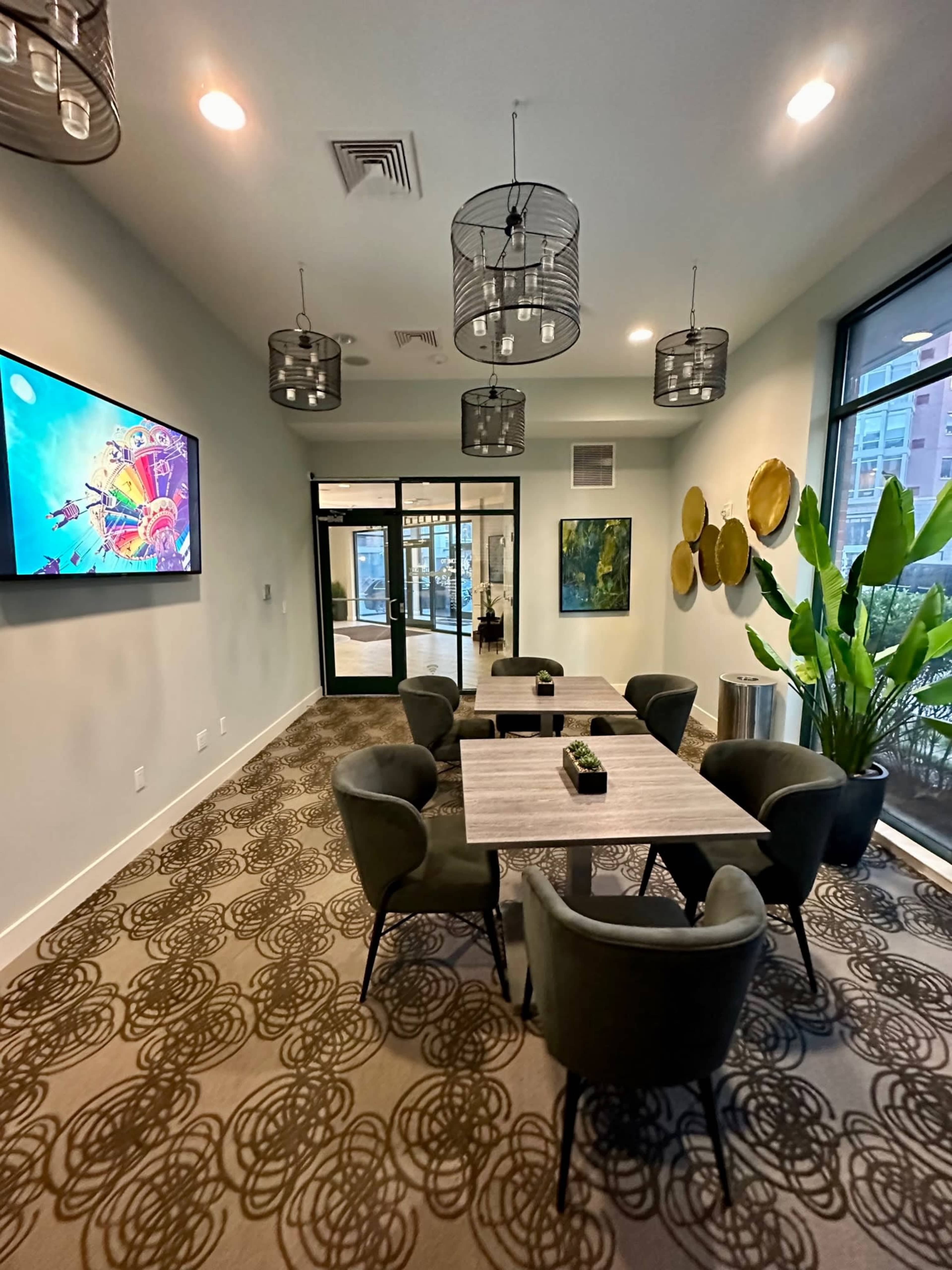 A well-lit meeting room features a large table surrounded by six chairs, with a television screen on the wall and greenery near the windows.