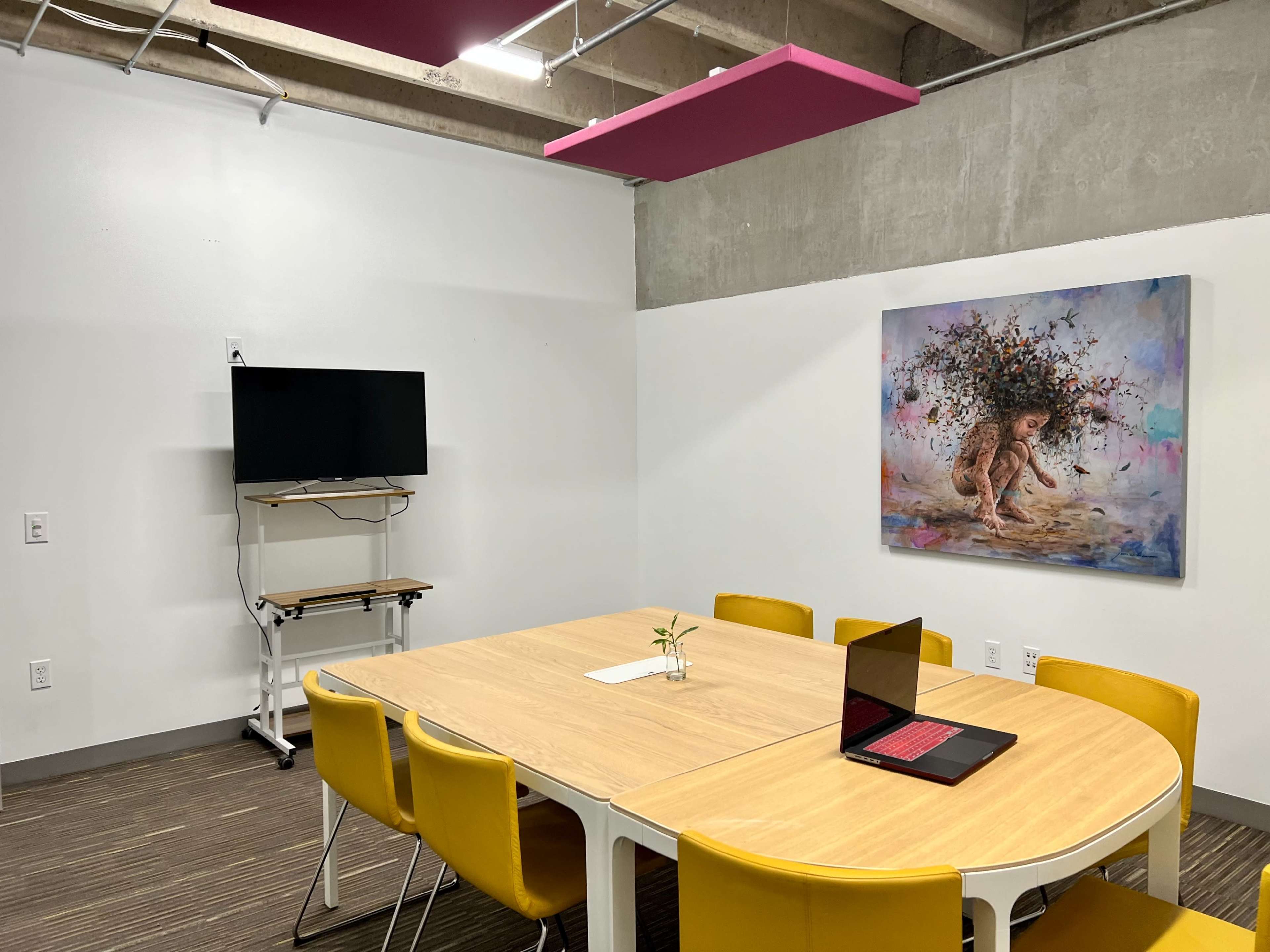 A meeting room features a wooden table surrounded by yellow chairs, a wall-mounted TV, and a colorful artwork depicting a lion.