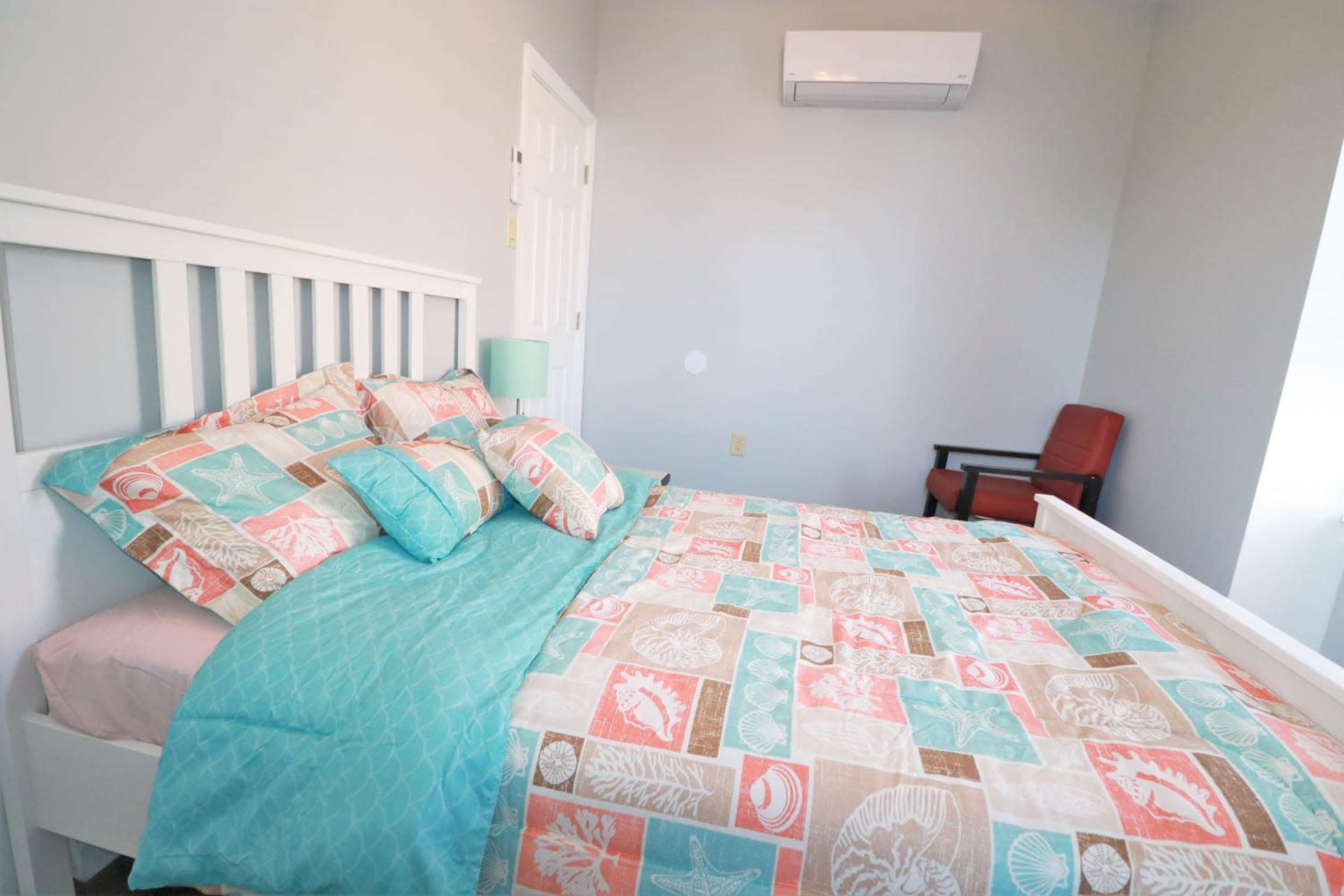 A neatly made bed with colorful patterned bedding is positioned in a softly lit bedroom, featuring a small side table and a chair in the corner.