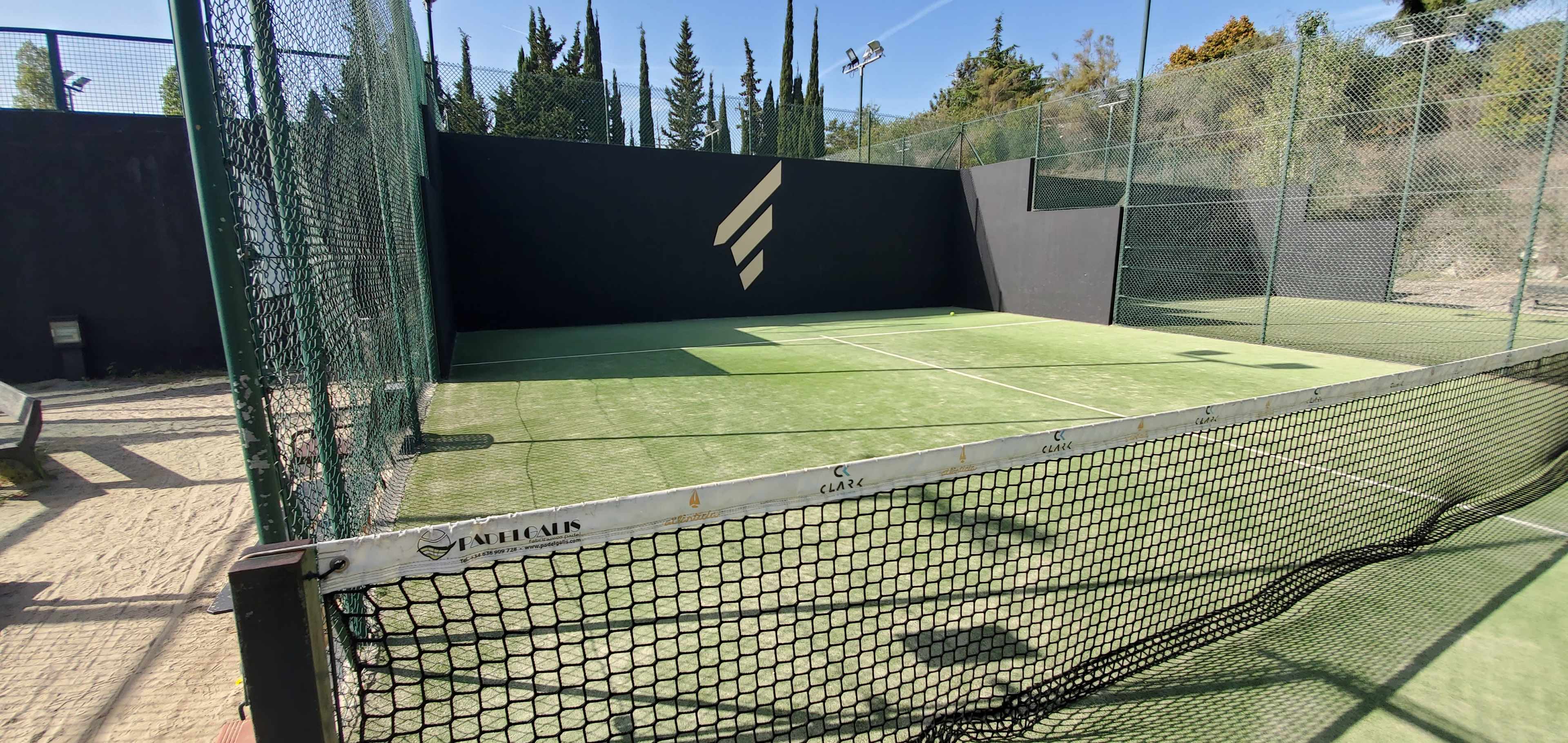 An enclosed outdoor padel court with a green artificial turf surface and a black wall featuring a logo.