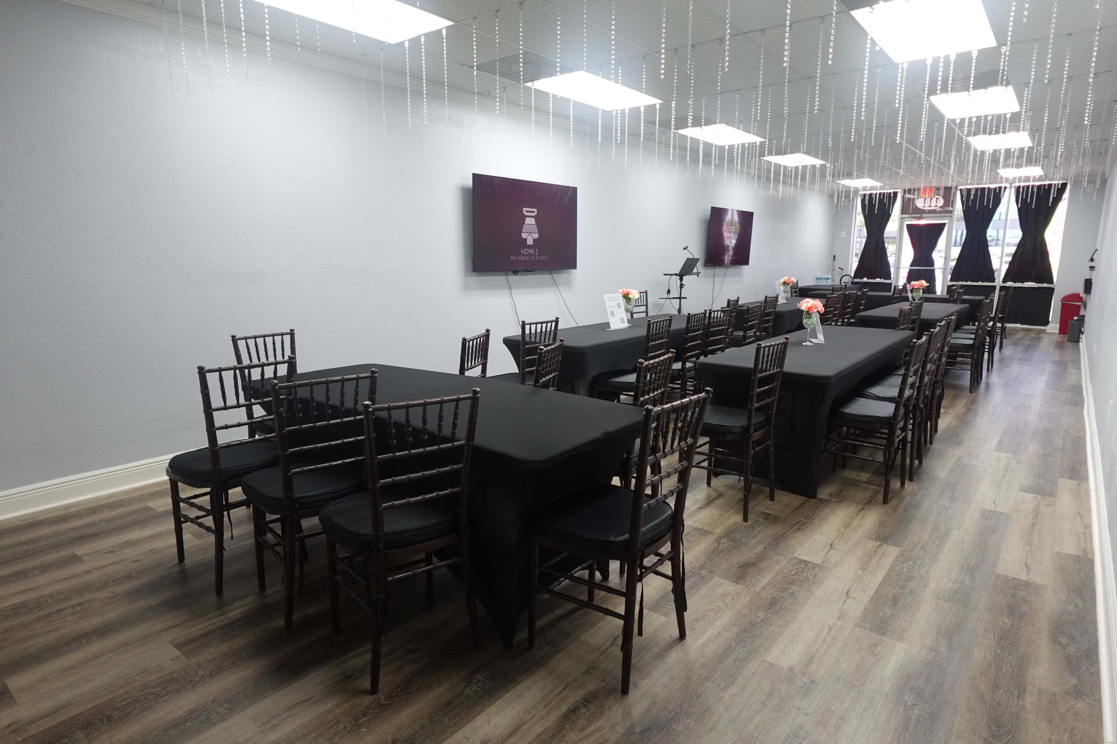 A long, narrow conference room with black tables and chairs, decorated with hanging string lights and floral centerpieces.
