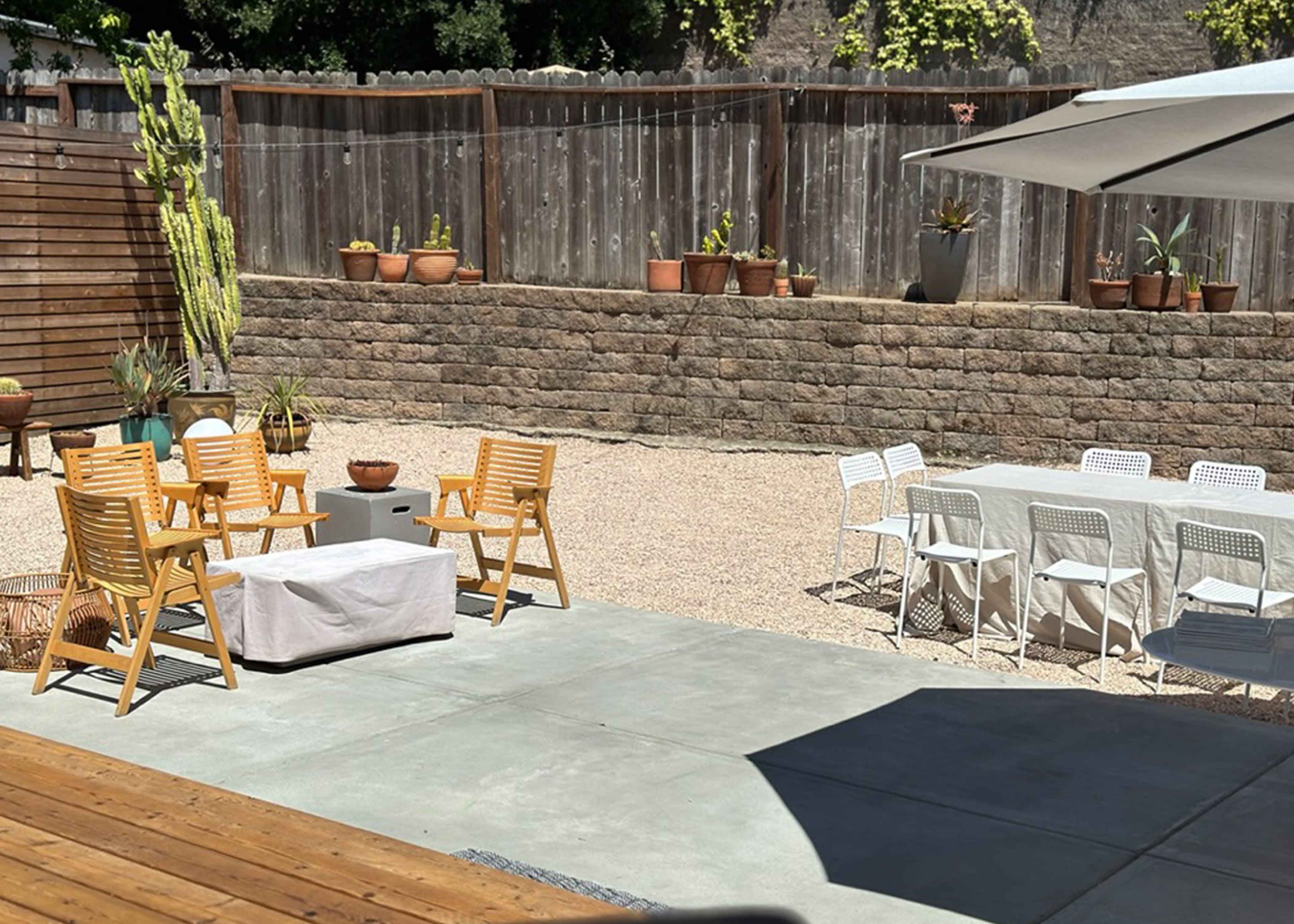 The scene depicts a spacious outdoor patio with wooden chairs, a table set for dining, and various potted plants along a stone wall.