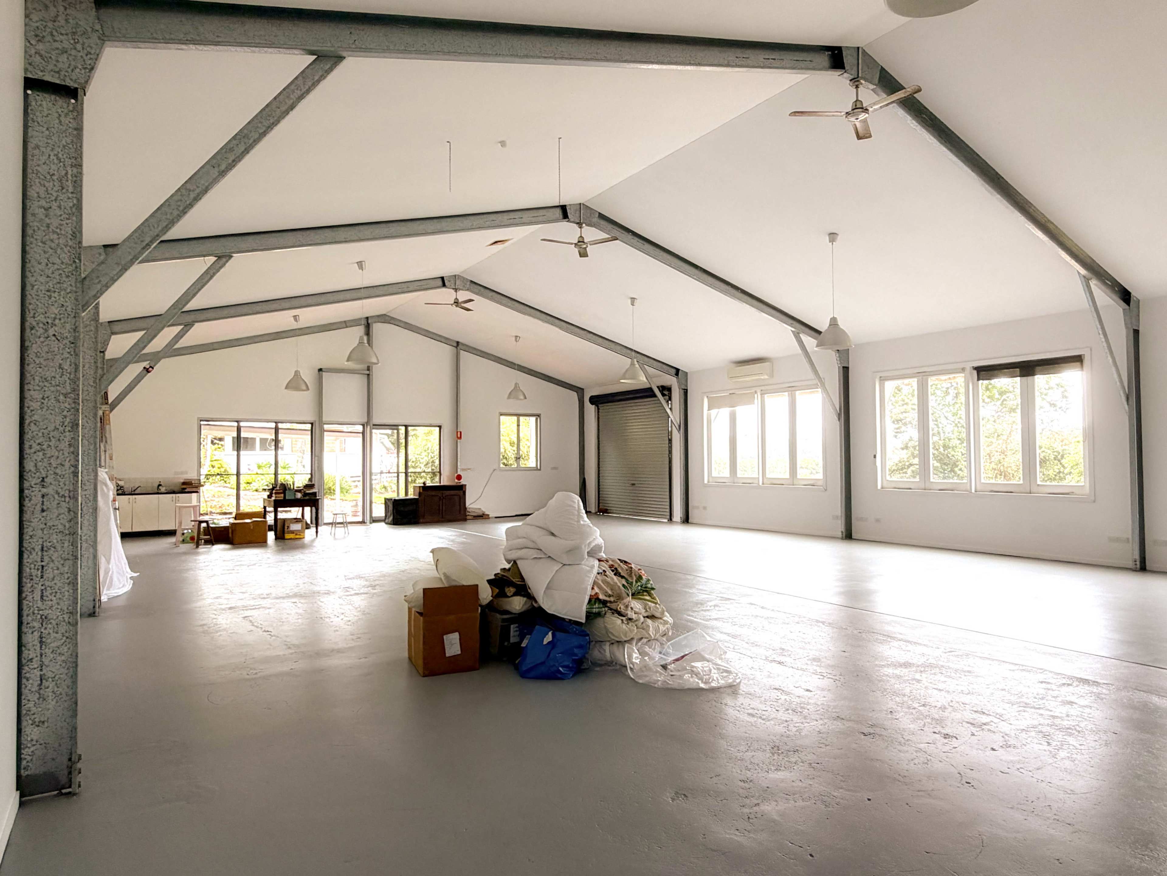 The image shows a spacious, empty room with high ceilings, metal beams, and a few scattered items such as boxes and a pile of fabric.
