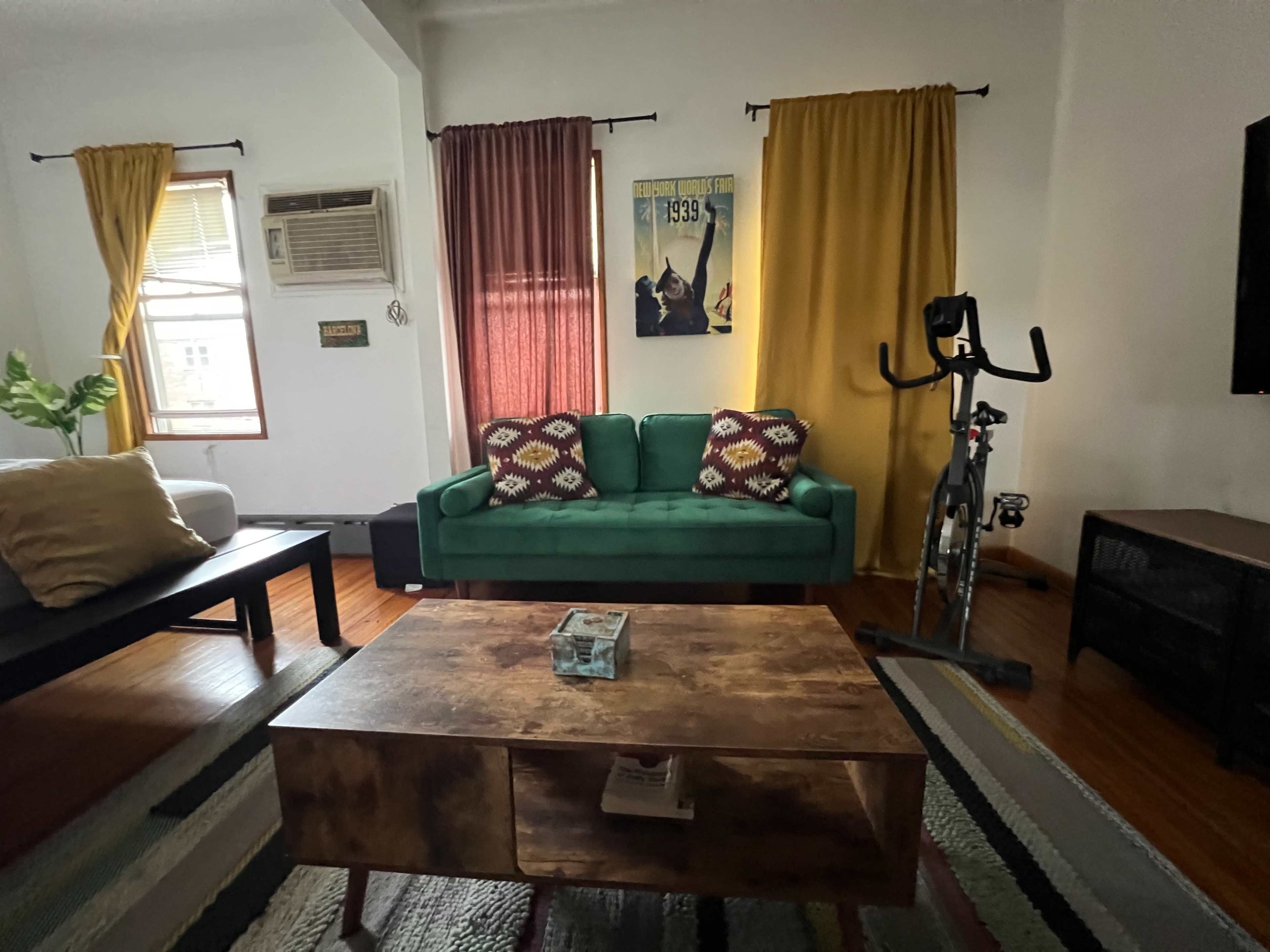 The living room features a green couch with patterned cushions, a coffee table, a bike on a stand, and curtains in shades of yellow and burgundy.