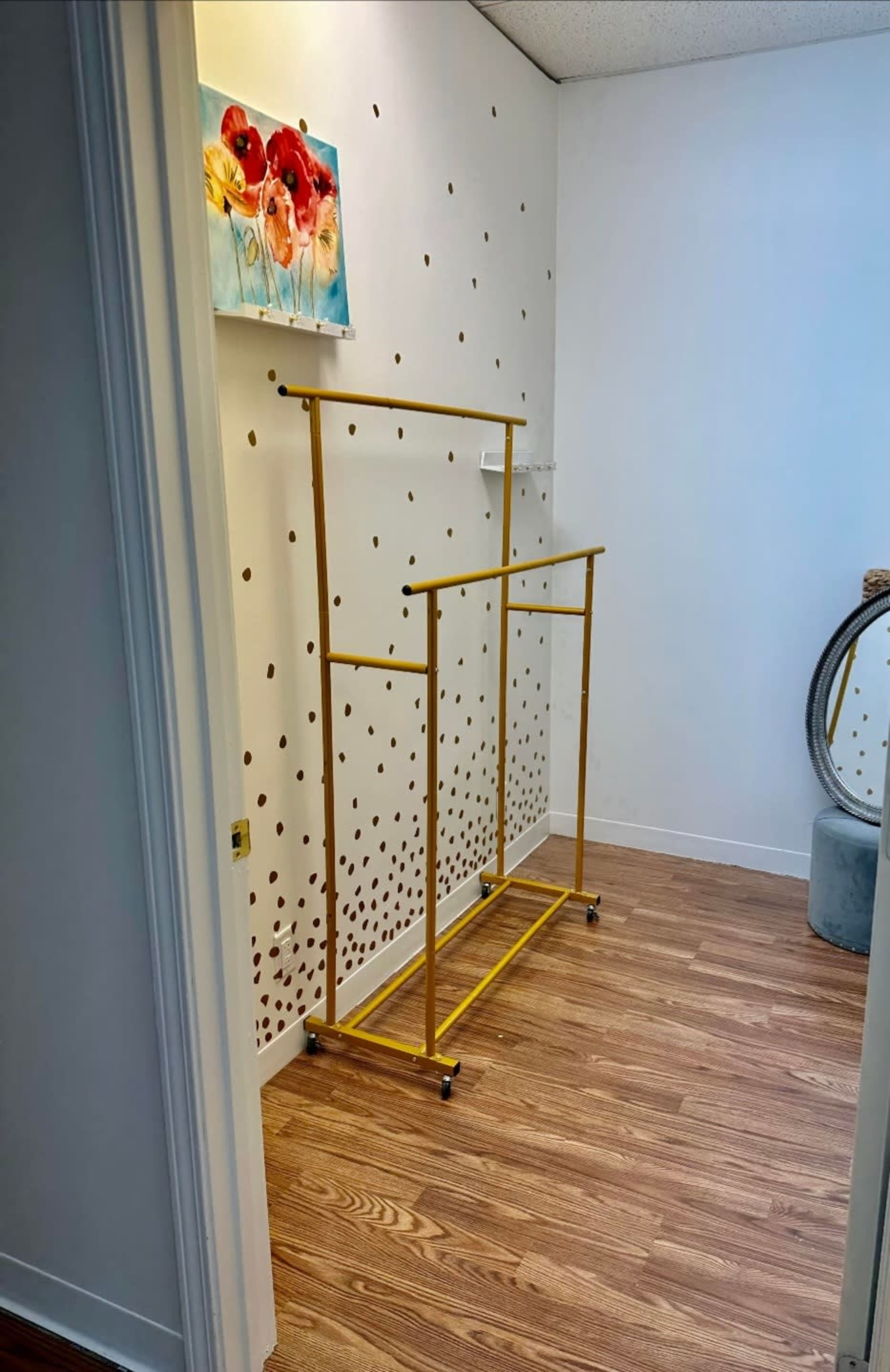 The image shows a brightly lit room with a wooden floor, featuring a yellow clothing rack, a circular mirror, and a floral painting on the wall.