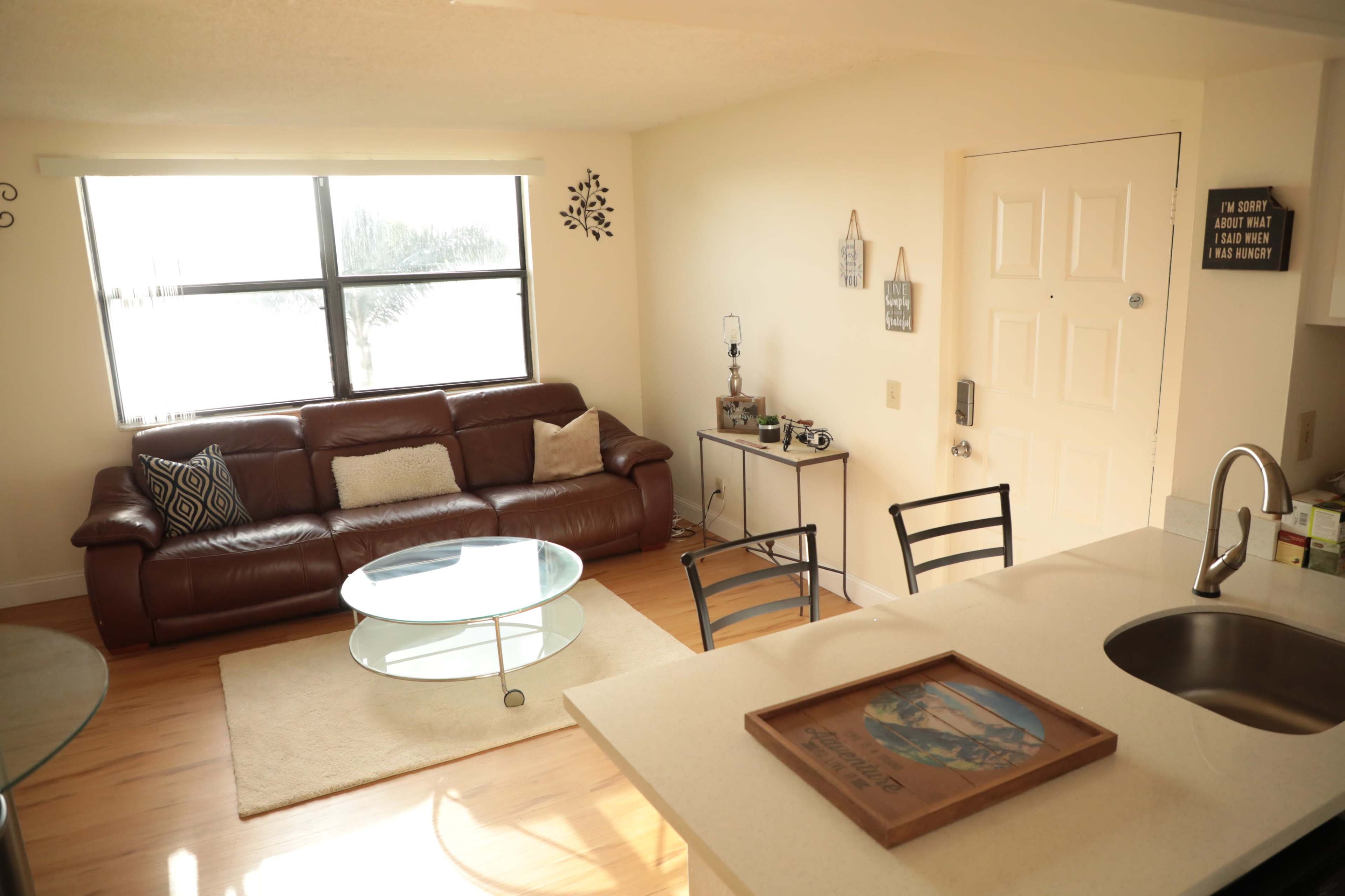 A living room with a brown leather sofa, a glass coffee table, and a small dining area with chairs, all illuminated by natural light from a window.