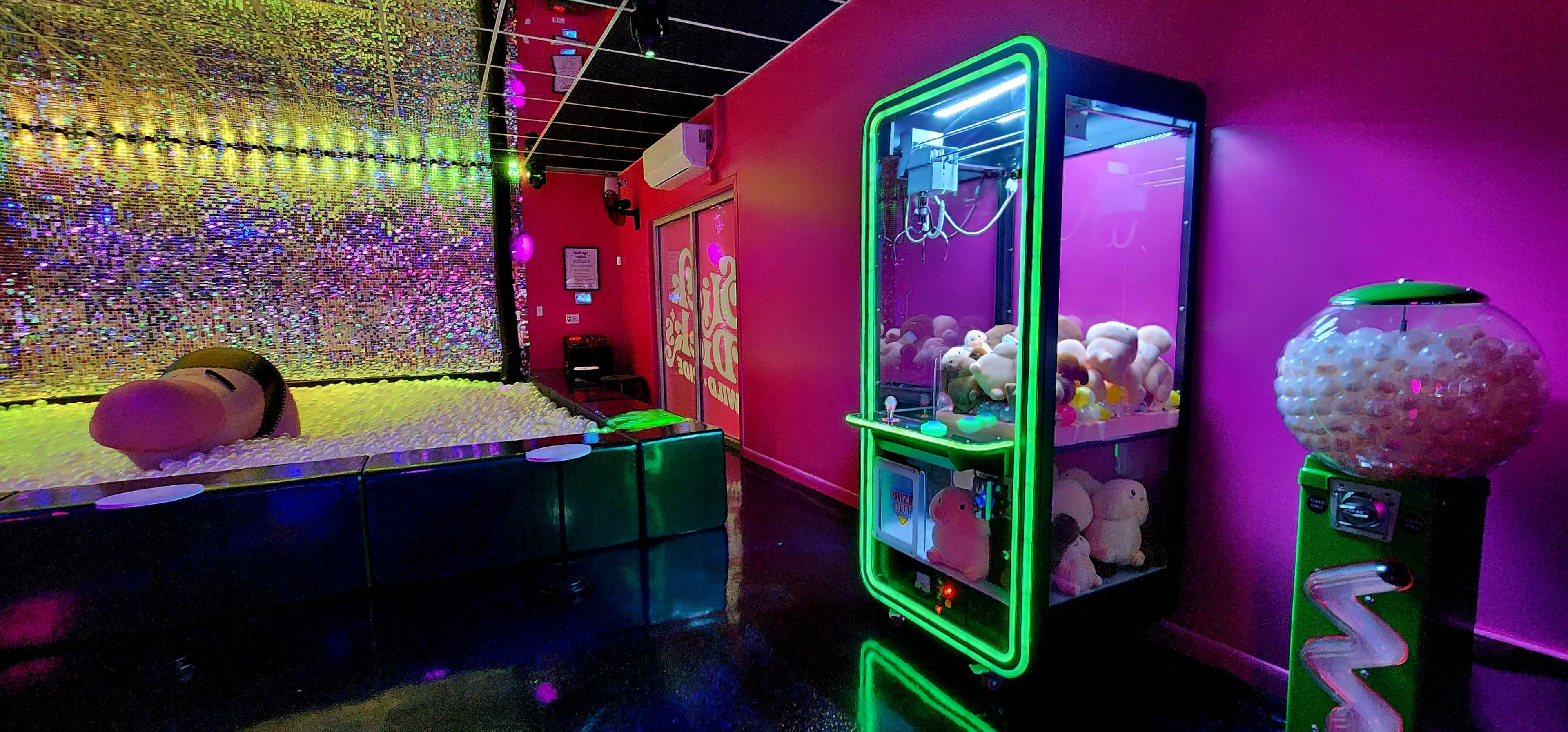 The image shows a vibrant room with a sparkling wall, a claw machine filled with plush toys, and a gumball machine, all against a backdrop of bright pink walls.