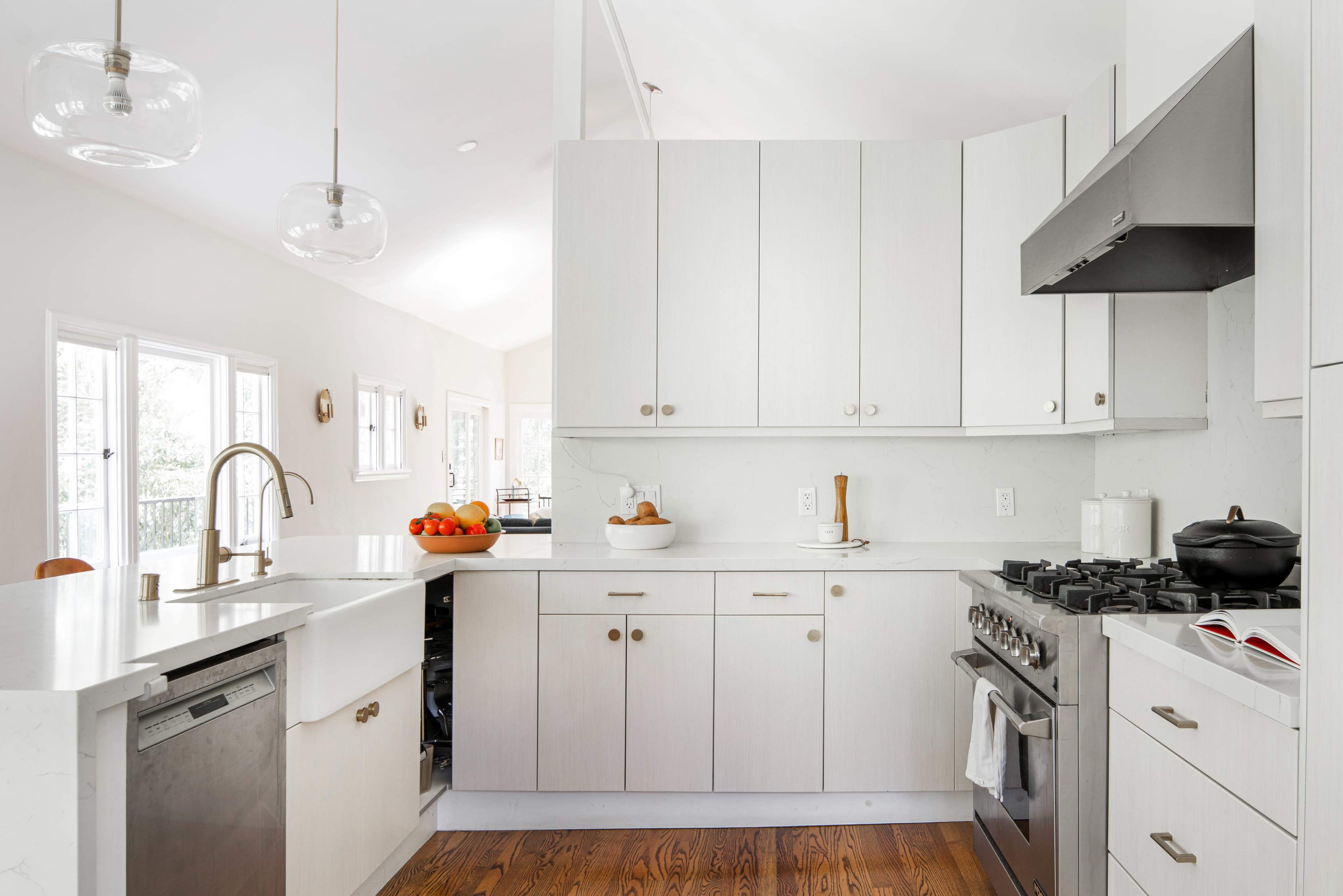 A modern kitchen features light wood cabinetry, a stainless steel stove, and a large sink with a gold faucet, complemented by large windows and decorative fruit.