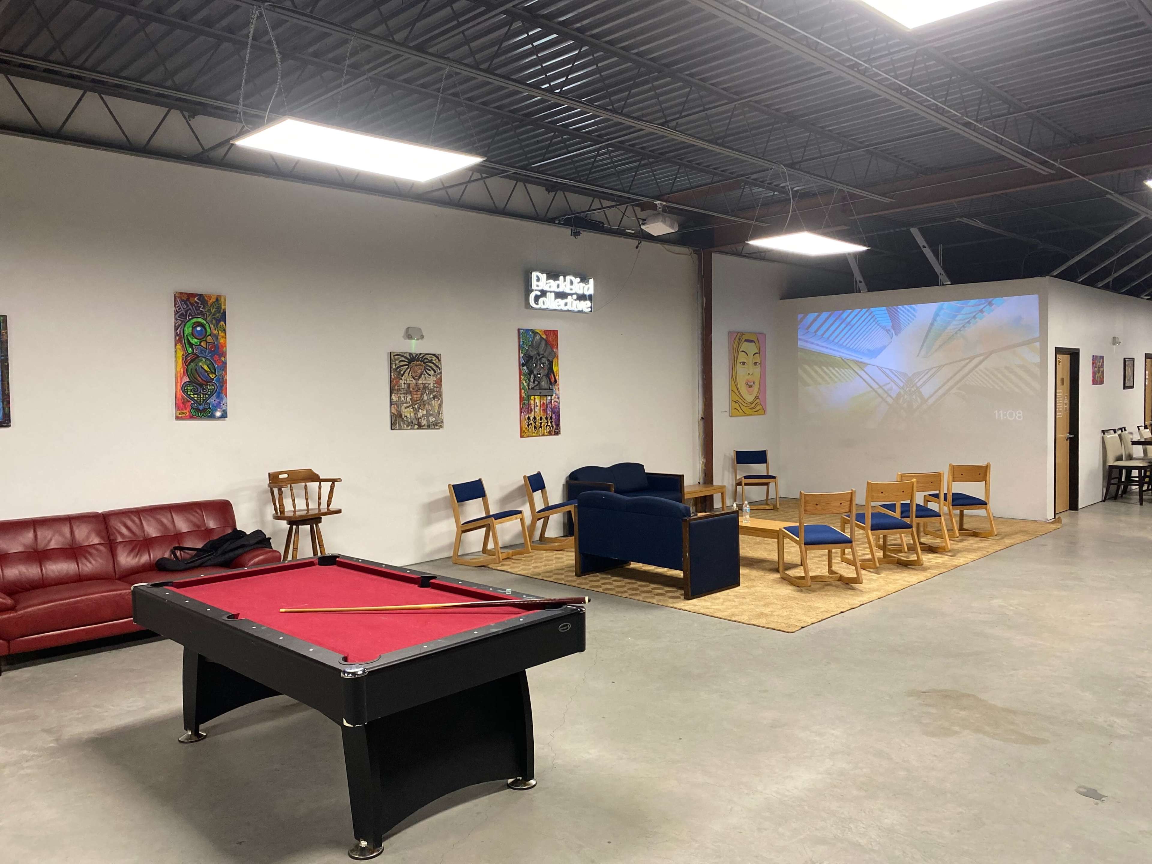 An indoor community space featuring a red pool table, a blue couch, several wooden chairs, and art displayed on the walls, with a projector screen showing a geometric design.