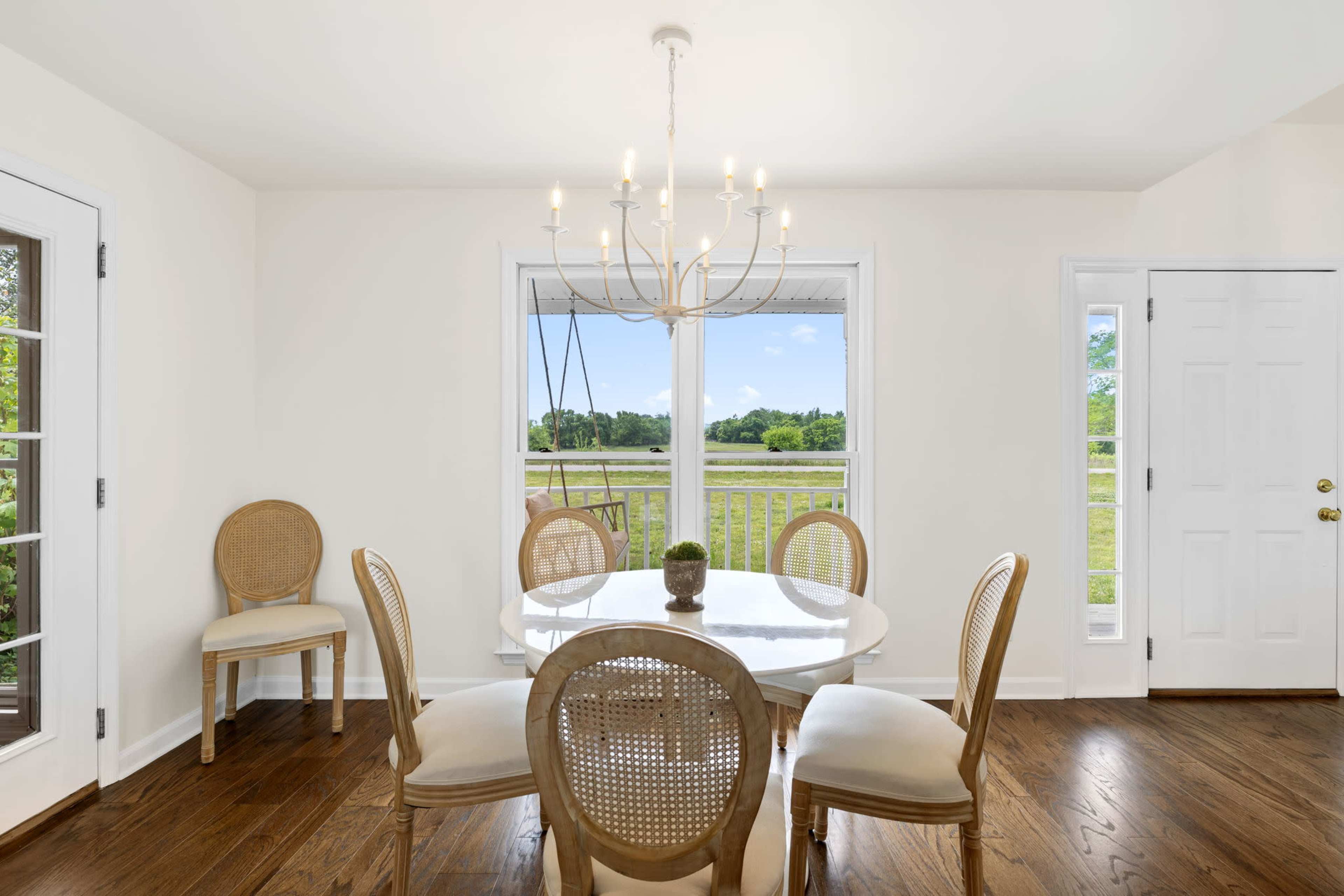 The image shows a well-lit dining area with a round table and six chairs, a chandelier overhead, and large windows overlooking a green outdoor landscape.