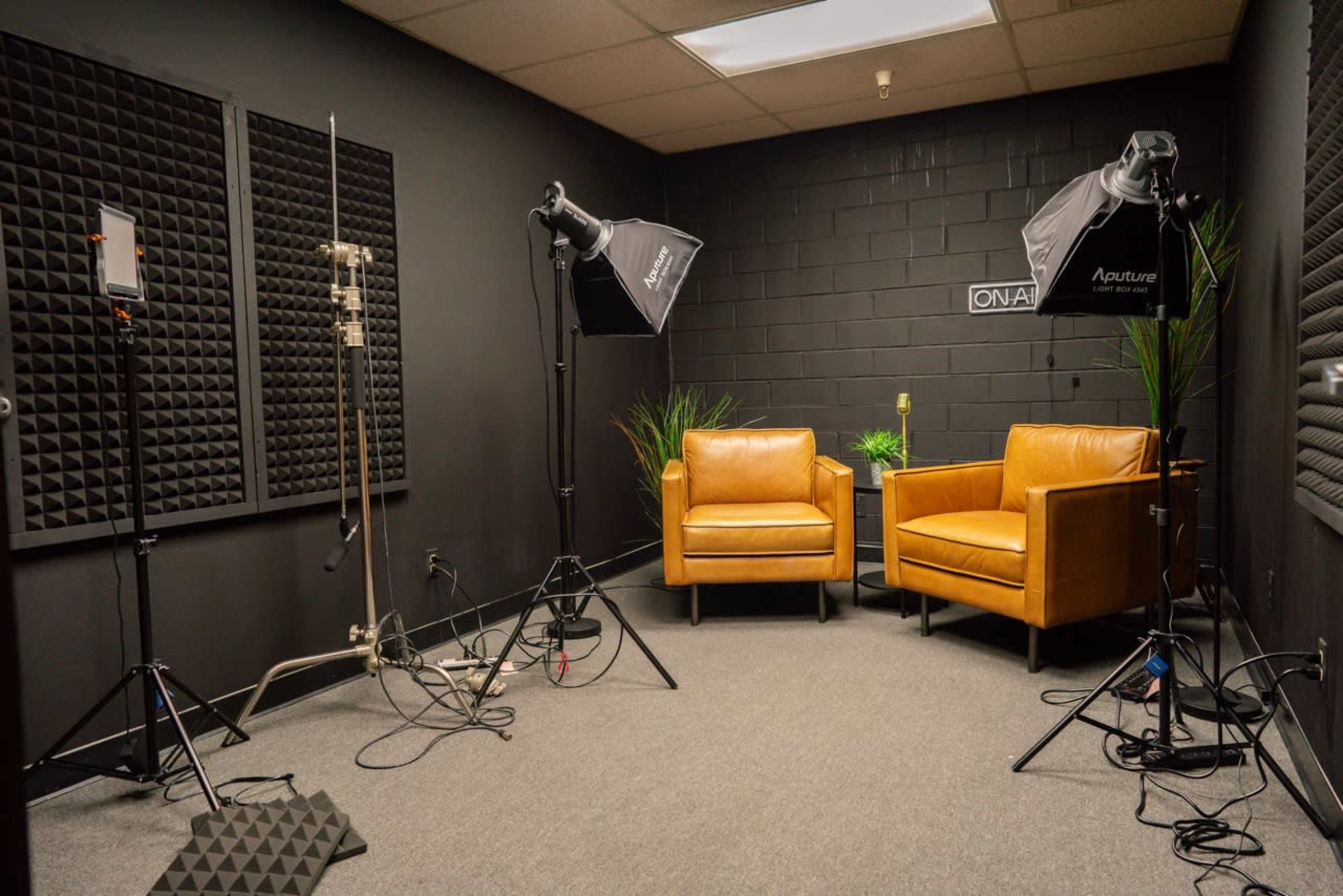 A small, dimly lit studio with two brown chairs, lighting equipment, and soundproof foam panels on the walls.