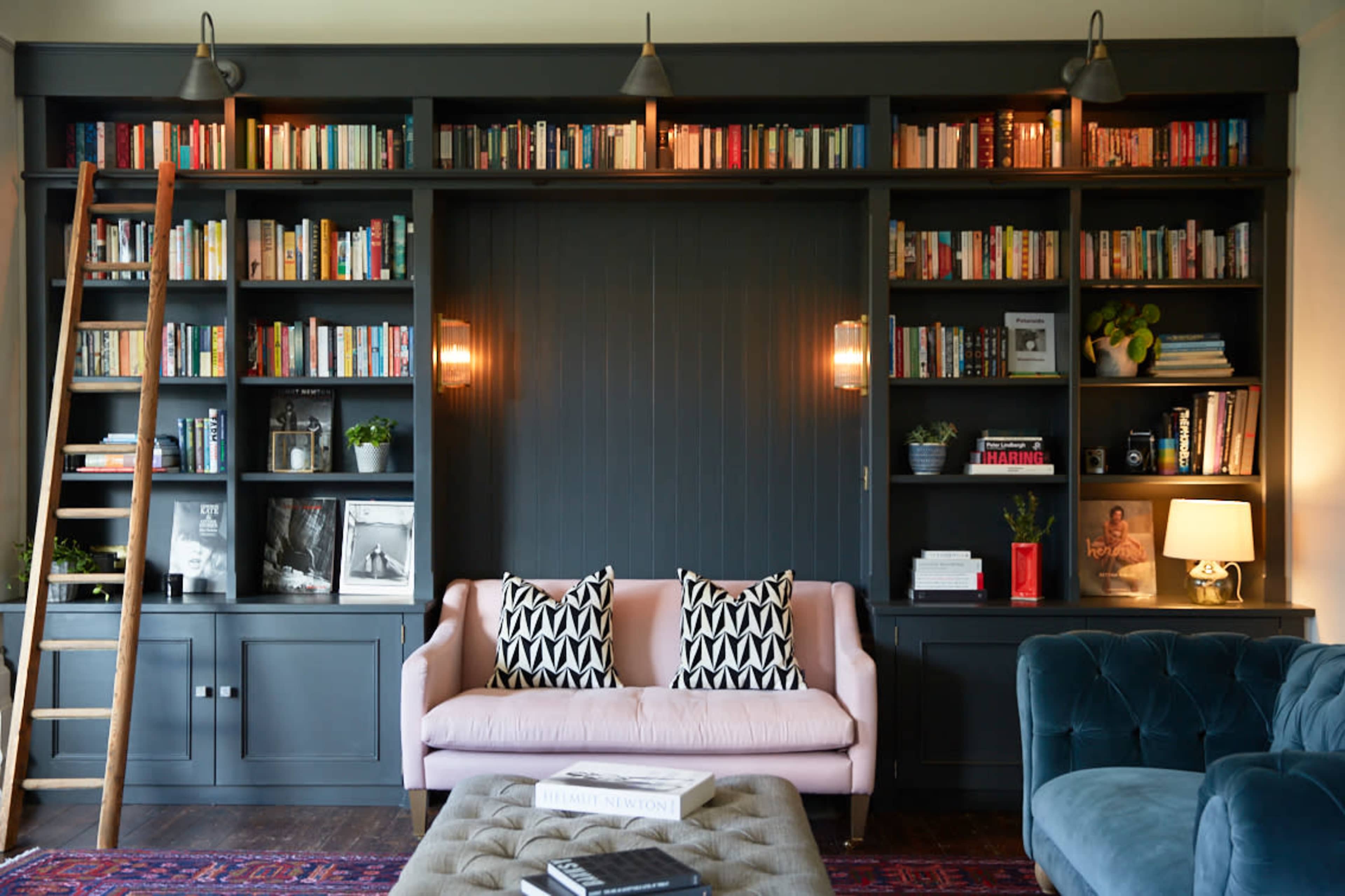 A modern living room features a large bookshelf filled with books, a pink sofa with patterned cushions, a blue velvet armchair, and warm lighting.