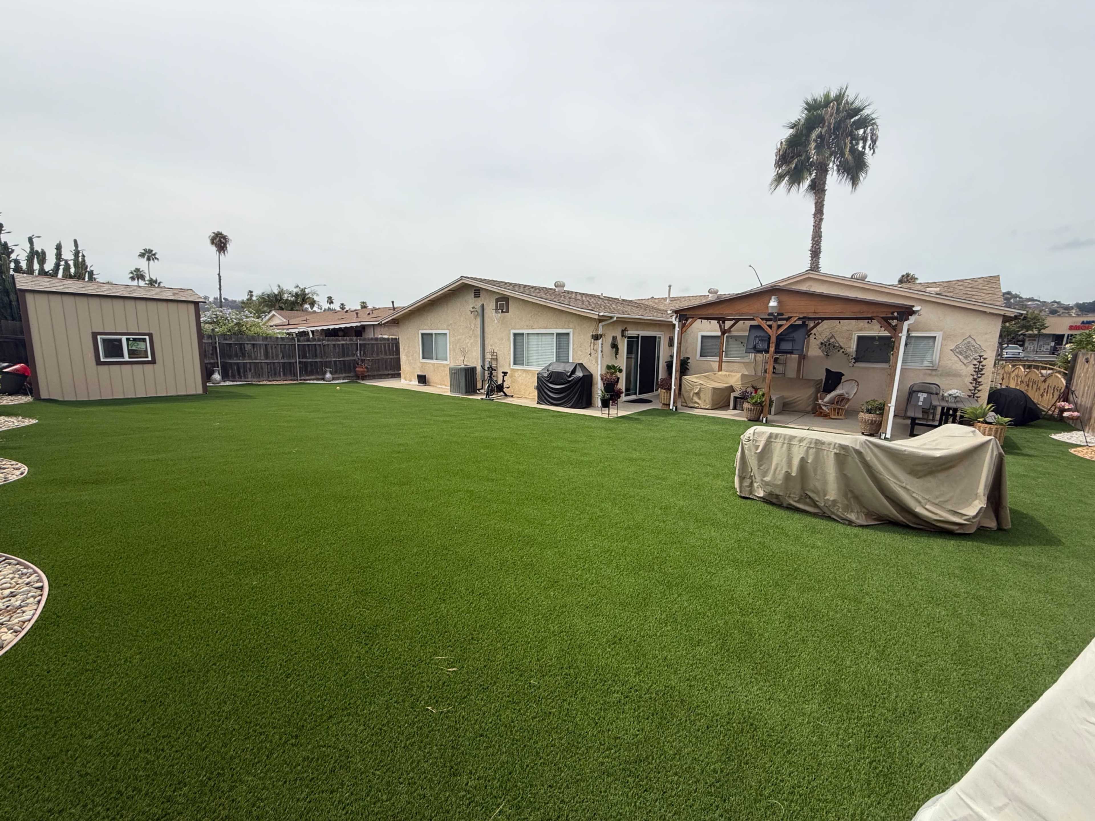 The image shows a backyard with artificial turf, a house with a porch, and outdoor furniture covered for protection.