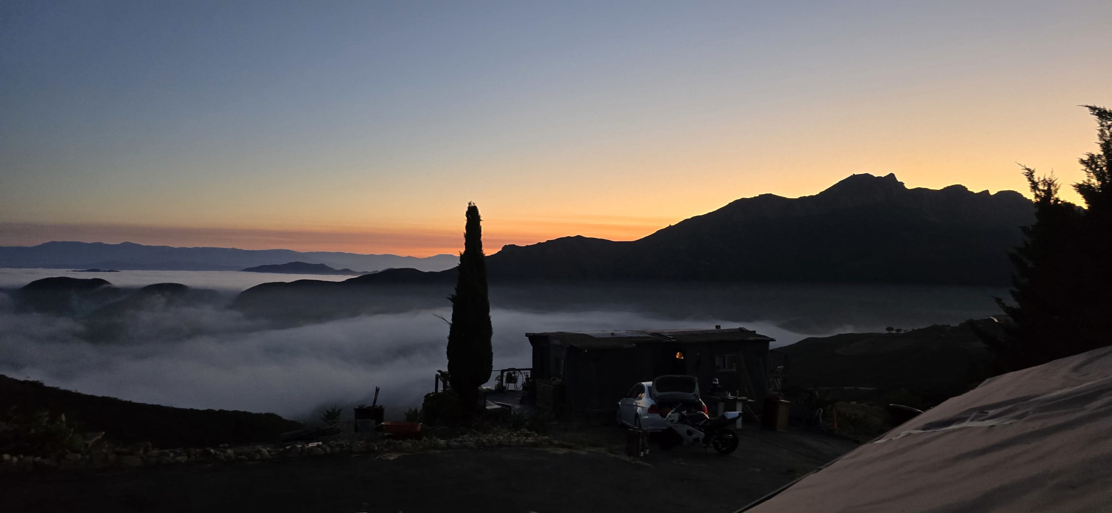 The image shows a mountain landscape at dawn, with rolling fog below and a silhouette of trees and a building in the foreground.
