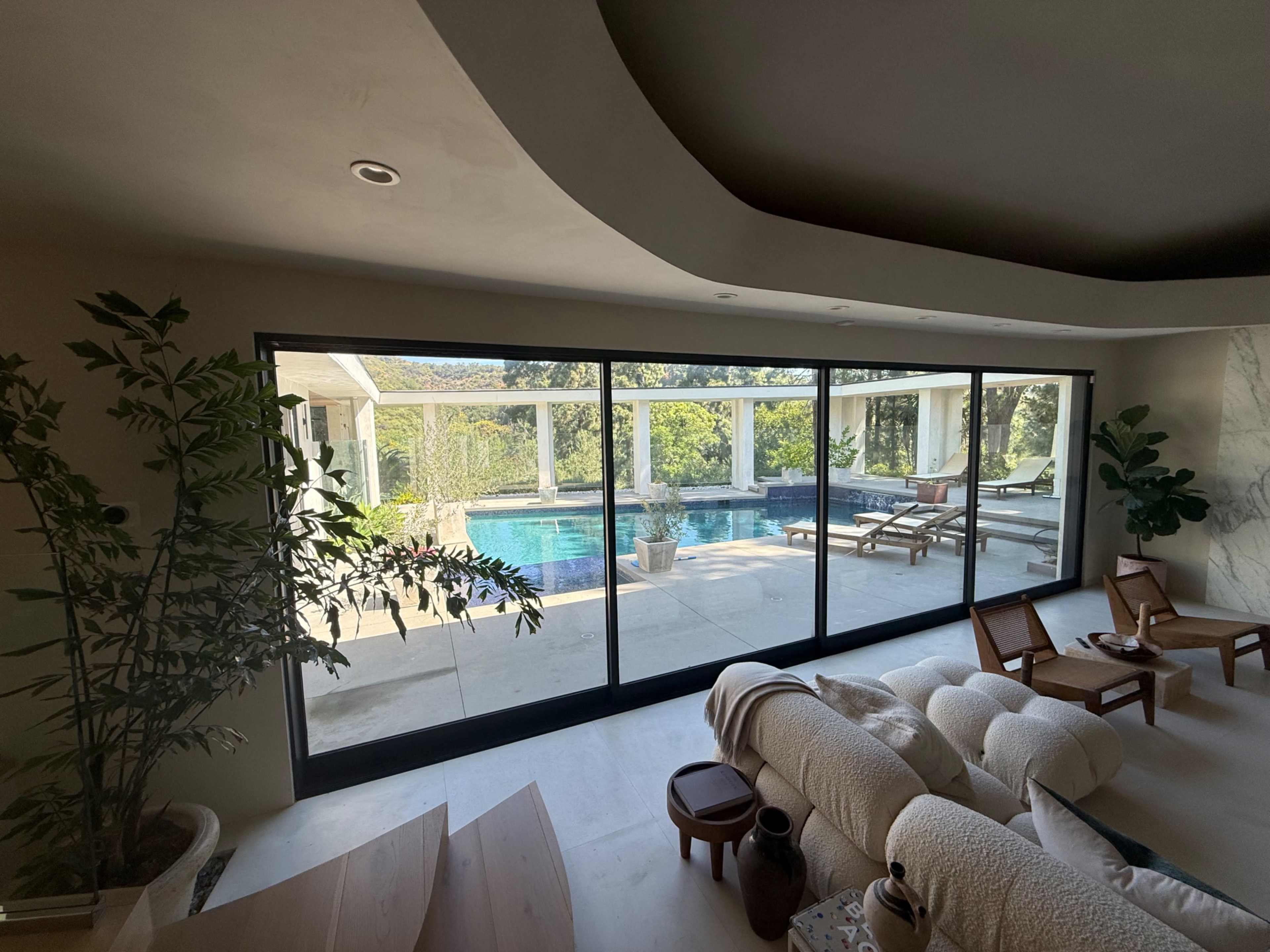 The image shows a modern interior with large glass doors that open to a backyard pool area surrounded by greenery and lounge chairs.